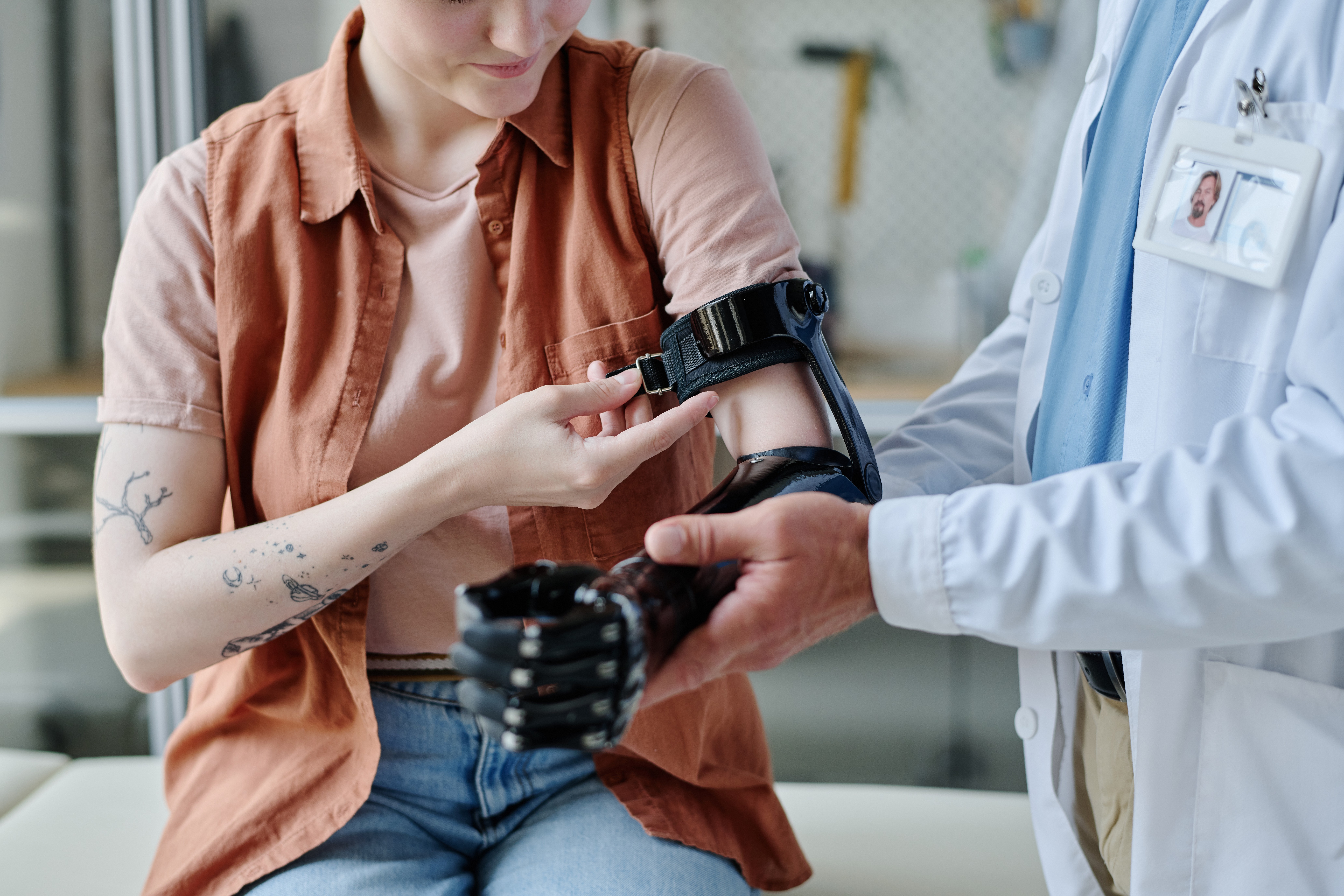 Clinician in a white coat fitting a black prosthetic arm and hand for a seated young woman in an orange shirt