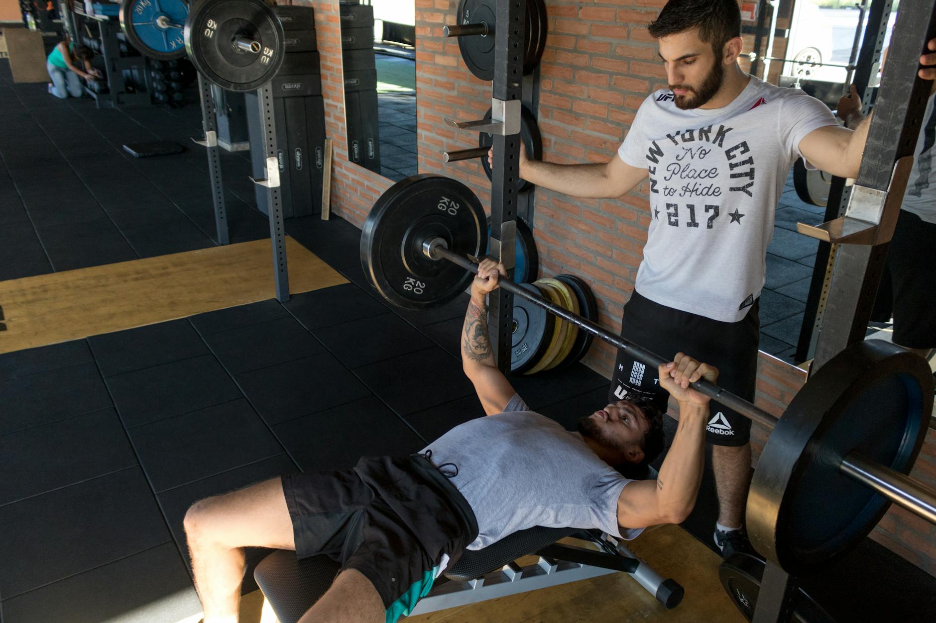 Man bench pressing with a spotter in a gym. Focused mood, New York City shirt, brick walls, and weight racks.