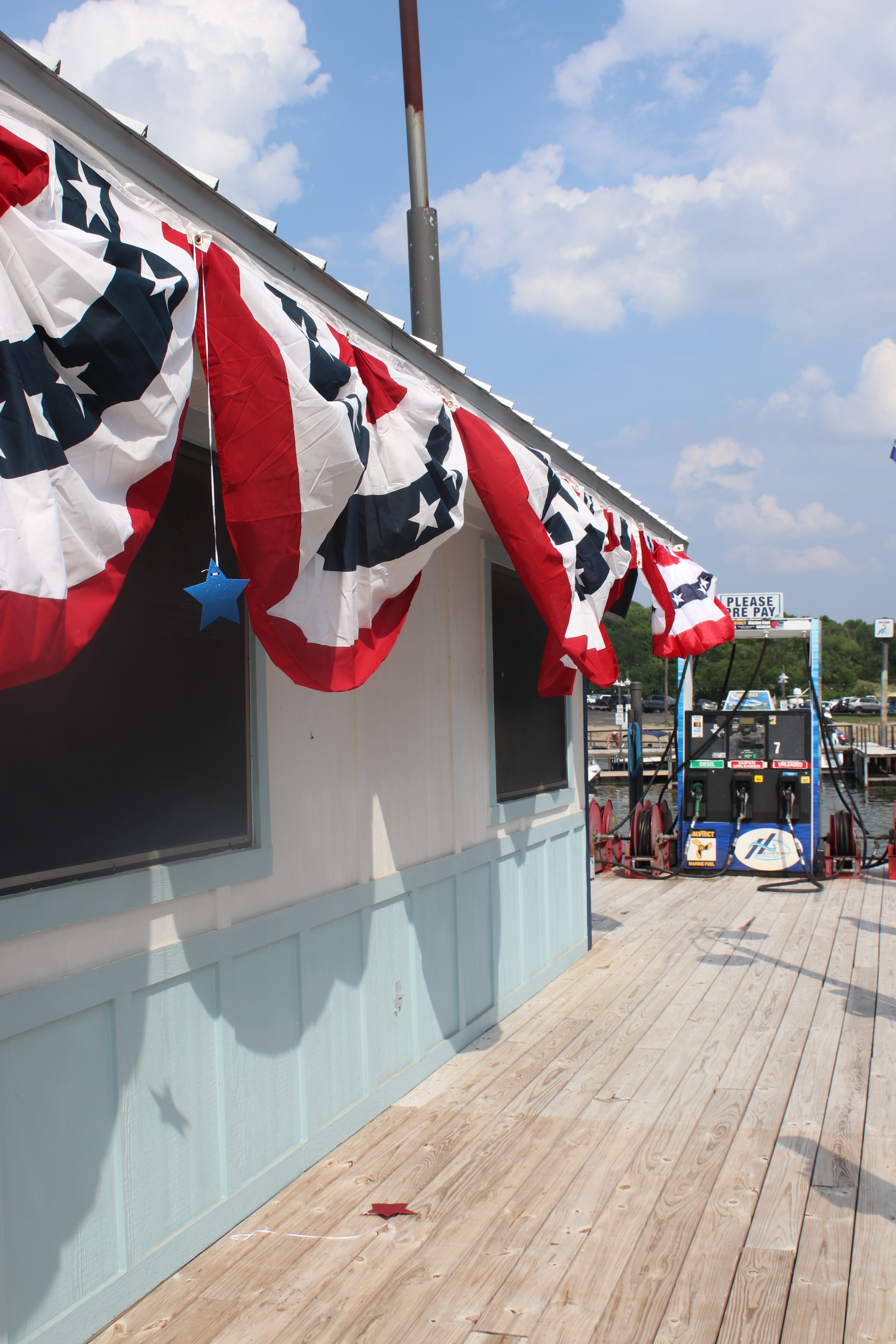 A dockside building is adorned with festive red, white, and blue bunting under a partly cloudy sky, with a view of an adjacent gasoline pump and the marina in the background.