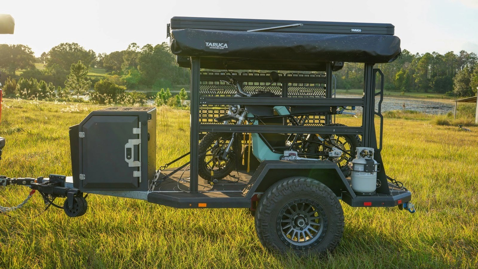 A utility trailer with a rooftop tent and bikes is parked on a grassy field, surrounded by trees under a clear sky, creating an adventurous, outdoor vibe.