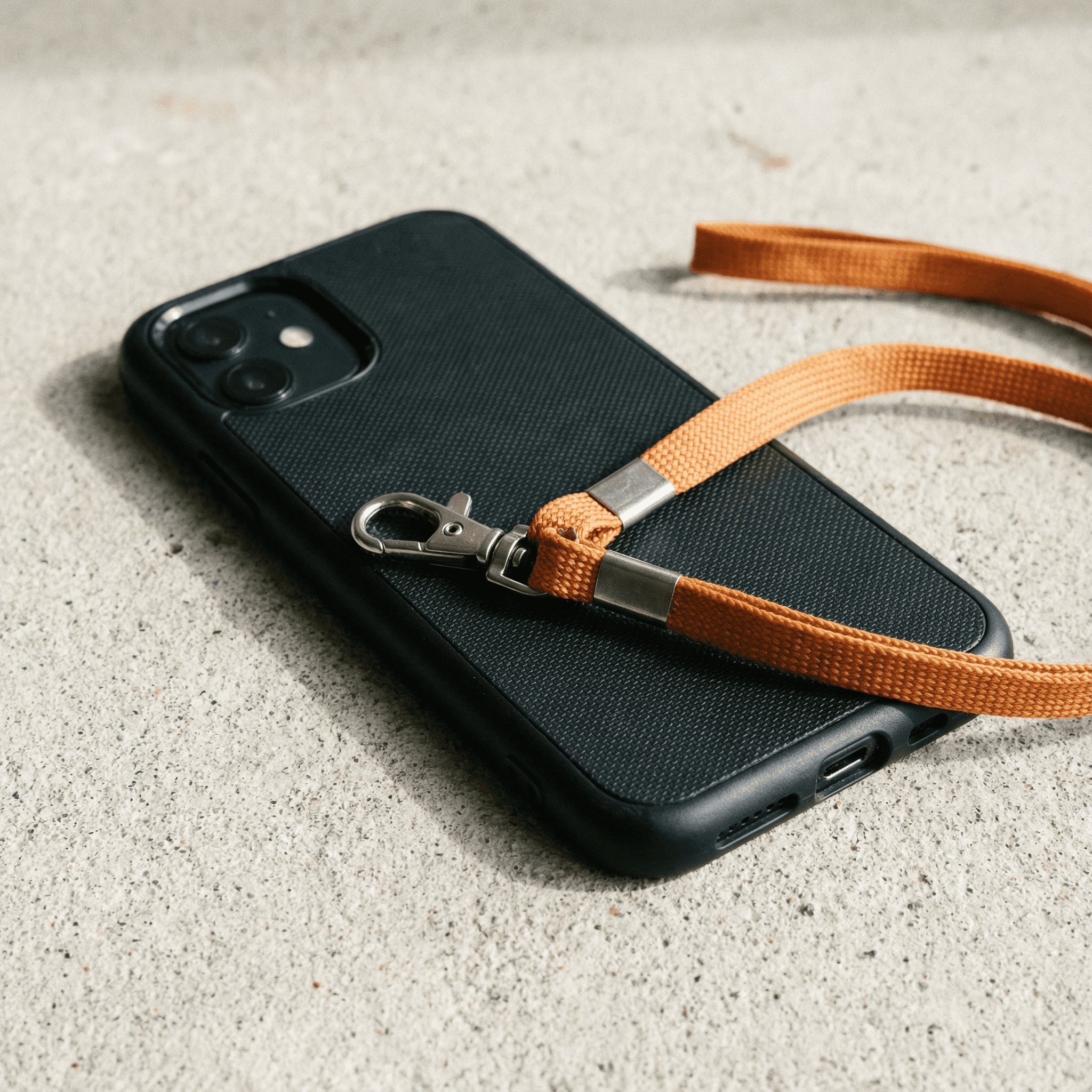 Black matte iPhone case lying flat on light surface, attached orange nylon wrist lanyard with metal clasp, product photography for accessories.