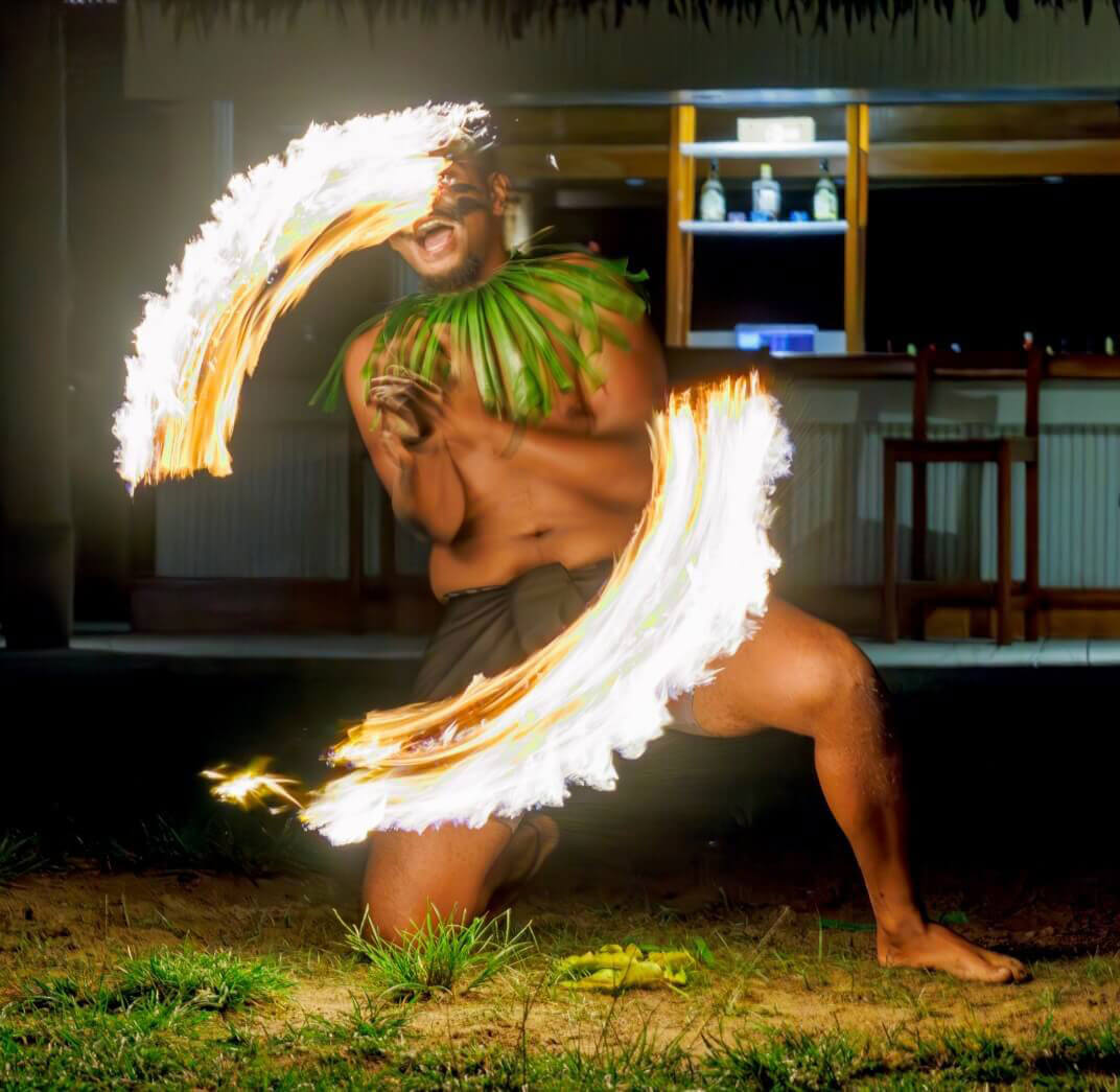 Energetic Fijian fire dancer in traditional attire at Uprising Beach Resort, spinning brilliant arcs of flame at night.