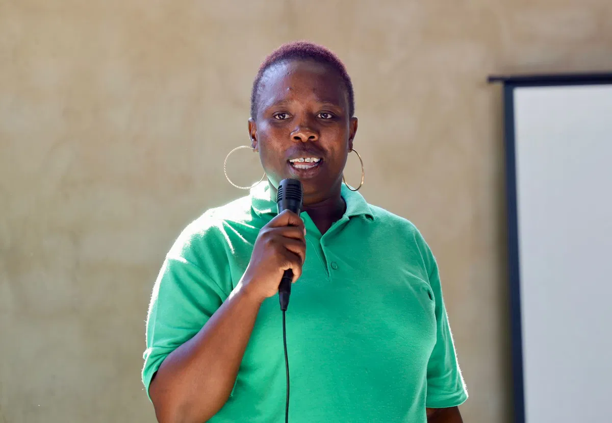 A smiling person with curly hair sits at a desk, looking at a laptop against a dark background.