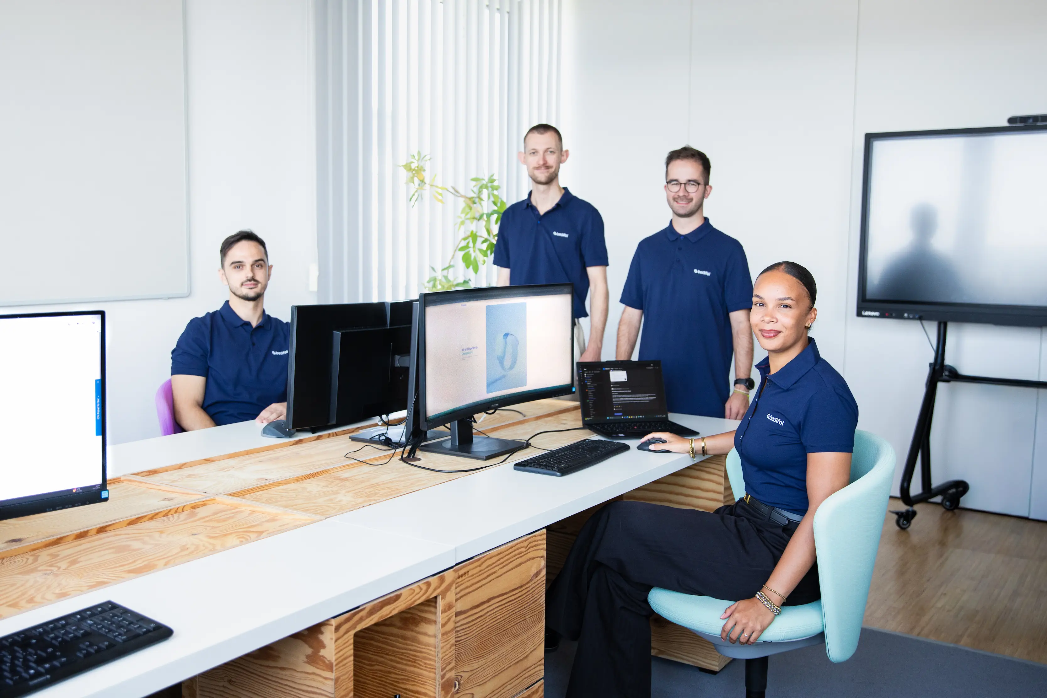 Four employees in Bedifol polo shirts at a desk with several monitors in the office.