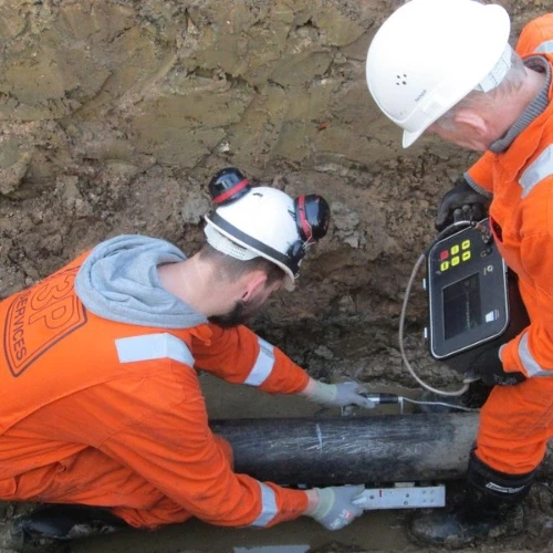 Two workers inspecting a pipe in a trench wearing safety gear, holding equipment for testing and maintenance.