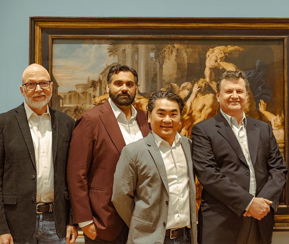 A group of four men in formal attire stand confidently in front of a large, classical painting, reflecting themes of art appreciation and networking within alternative investments.