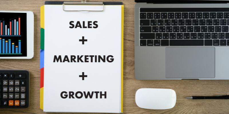 Sales plus marketing plus growth plan on a clipboard next to a calculator and laptop