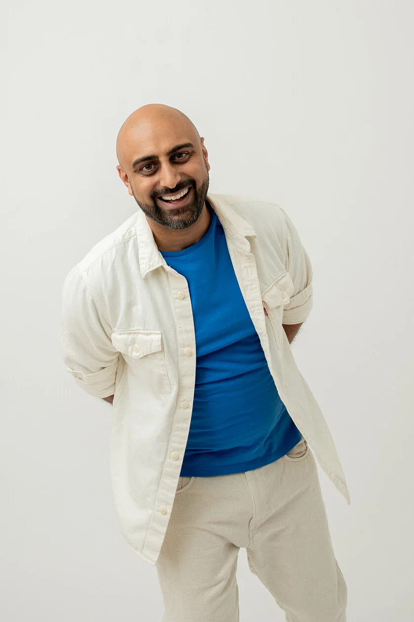 Smiling man in blue shirt and cream button-up jacket with hands behind back leaning slightly forward with a white background.