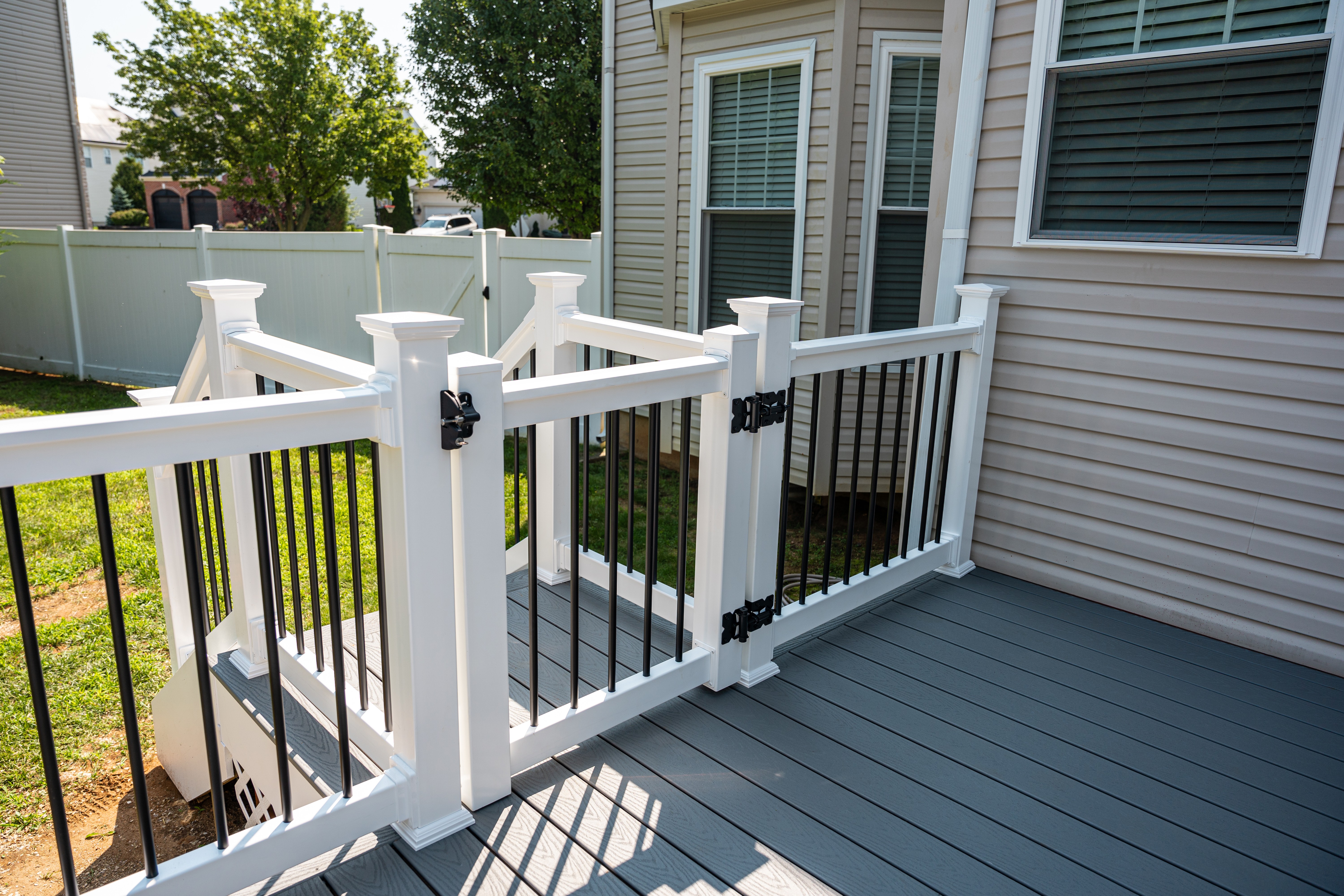 A spacious, modern backyard deck featuring gray composite decking with a white railing and black balusters, positioned beside a beige house with large windows, overlooks a green lawn enclosed by a white privacy fence.