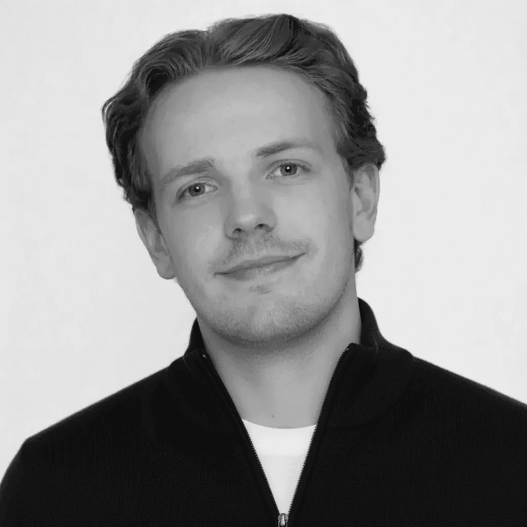 Young man with wavy hair wears a dark overcoat, rust-colored sweater, white shirt, and patterned tie, exuding a poised and sophisticated demeanor.