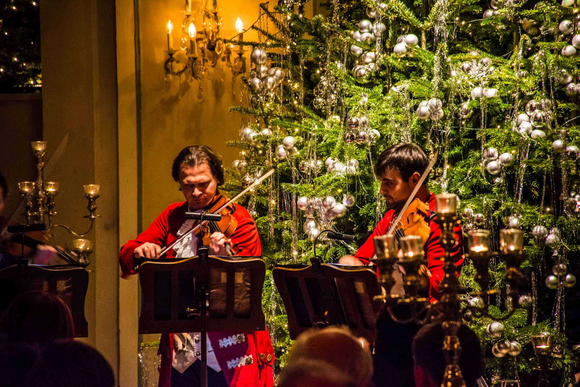 Two musicians in red shirts play accordions and a trumpet in a room decorated with green tinsel and white lights.