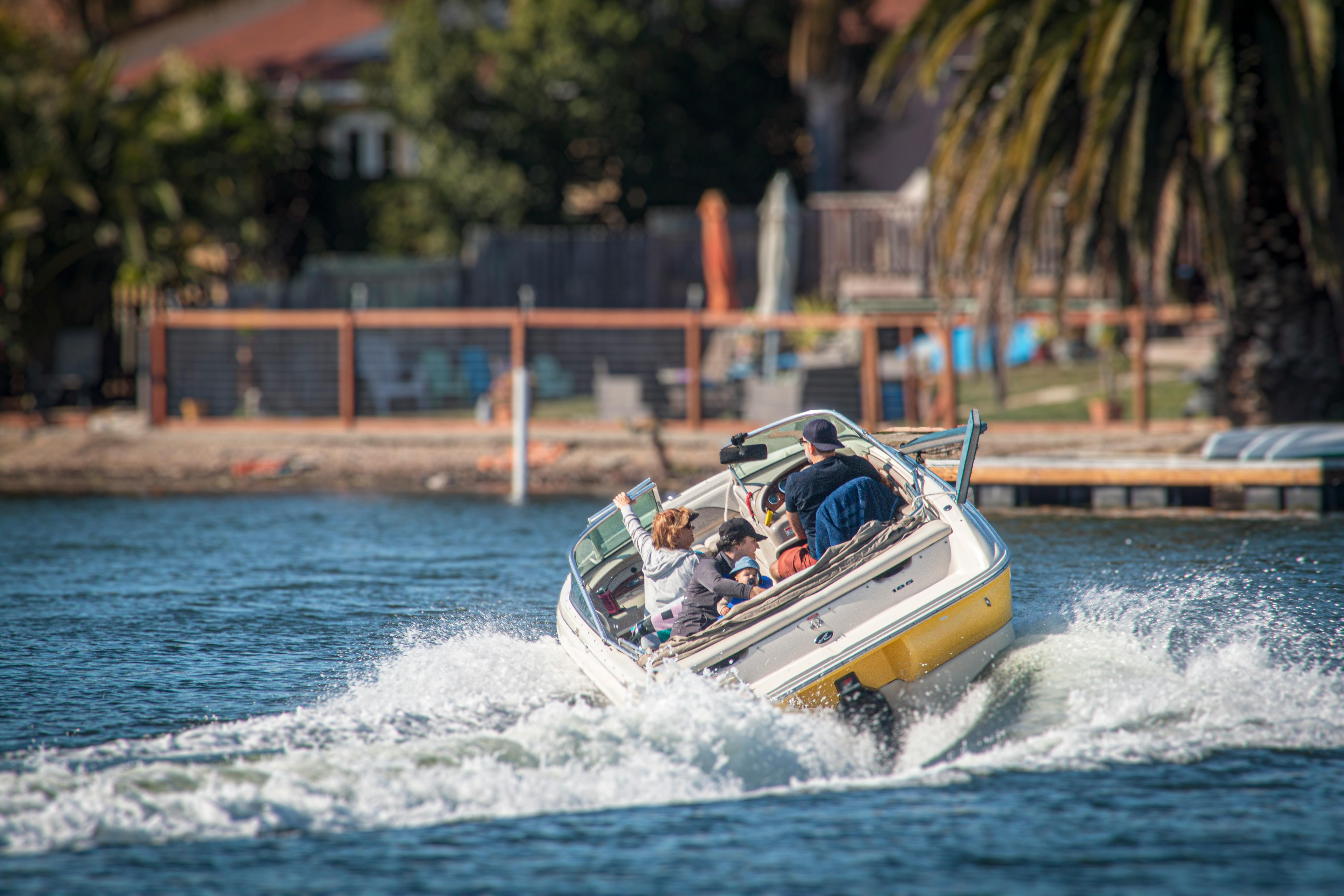 A small motorboat with people on board speeds through a calm lake, creating a trail of white waves, with a scenic background of palm trees and a distant shoreline.