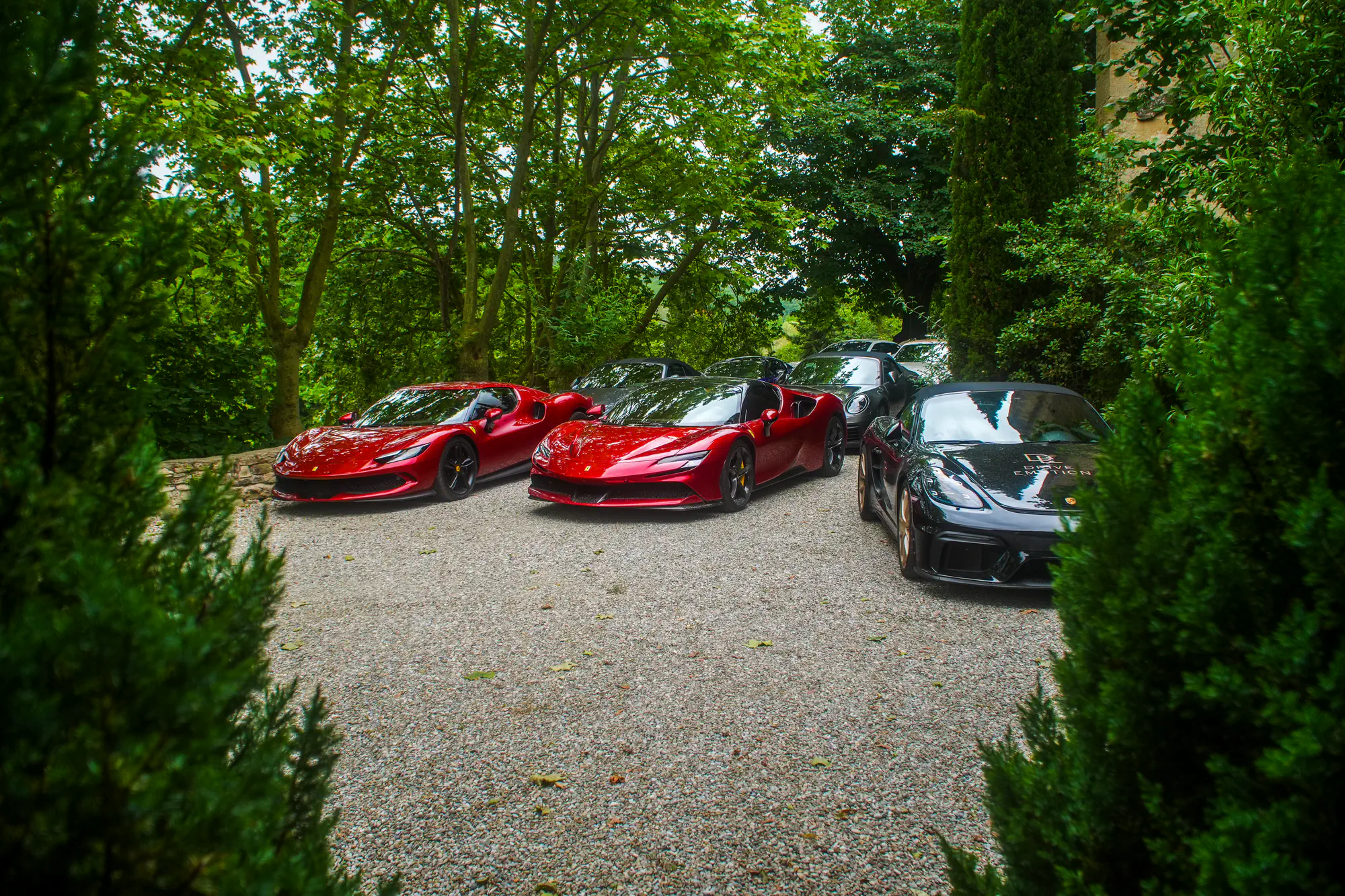 Deux Ferrari rouges et une Porsche 718 noire au logo Drive Emotion garées sur une cour en gravier, entre les arbres
