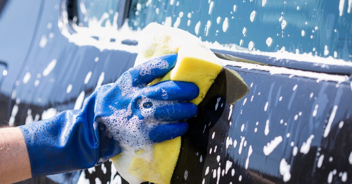 A close-up of a blue-gloved hand pressing a soapy, yellow sponge against a car window and door outside.