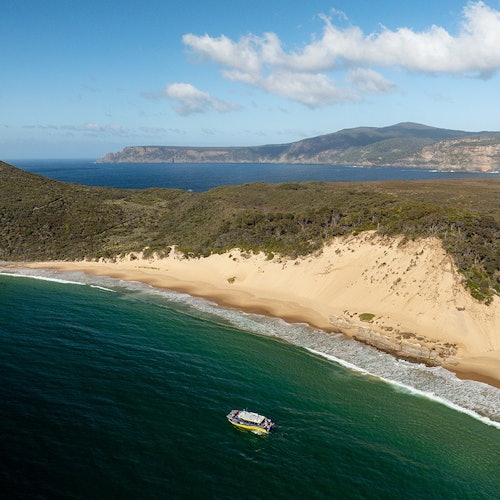 從空中俯瞰，看到一艘船在綠色的水面上，旁邊是沙灘，背後是崎嶇的山丘和遠處的山脈，天空中雲層稀疏。