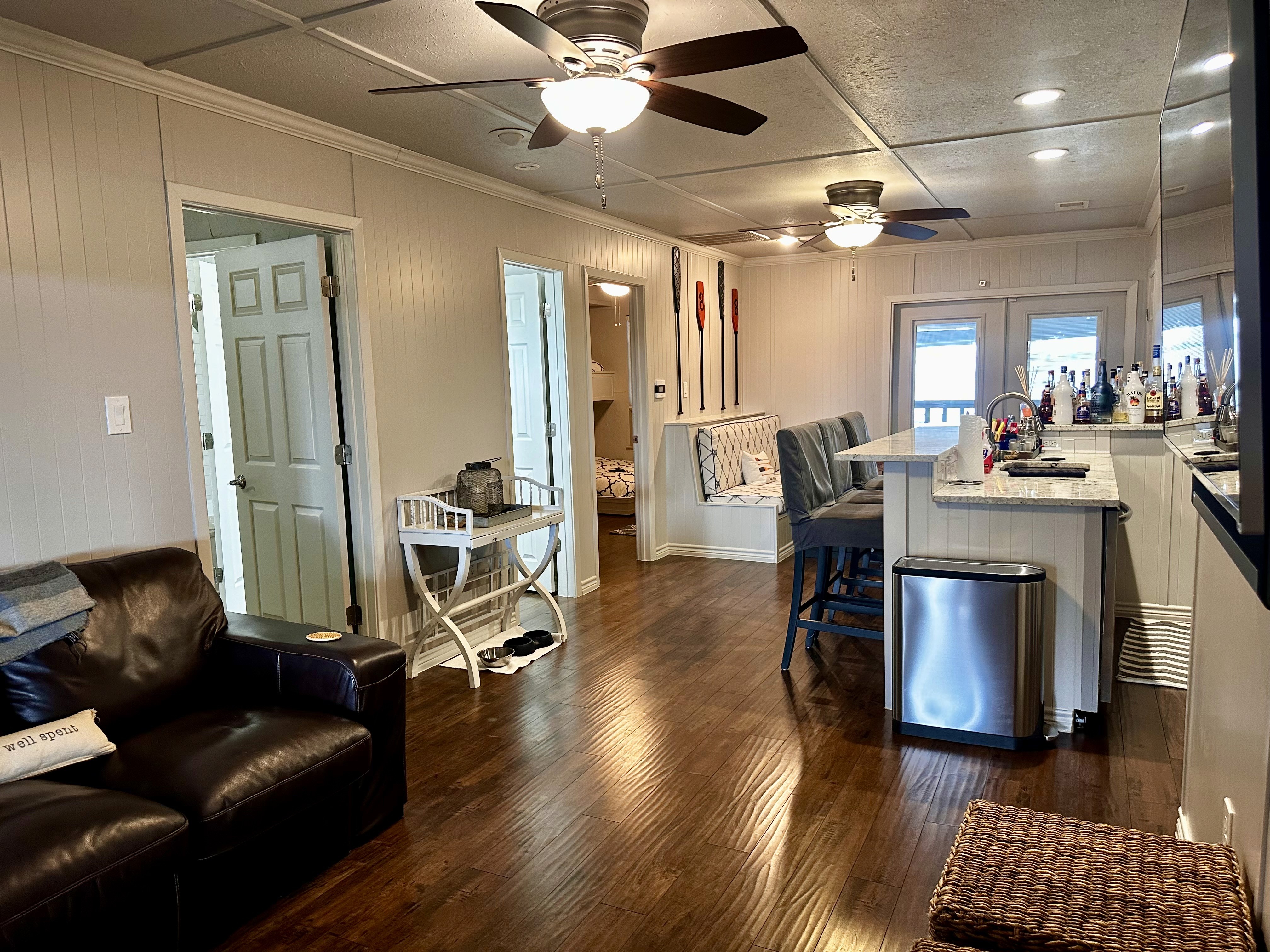 Interior shot of a living space with a kitchen, seating area, and doors. Hardwood floors and ceiling fans.