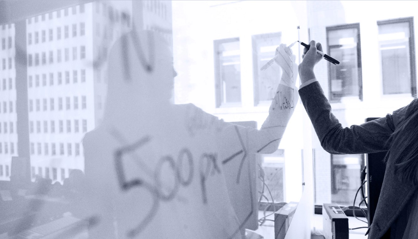 Photo of a woman in a suit writing on a white board in front of a window.