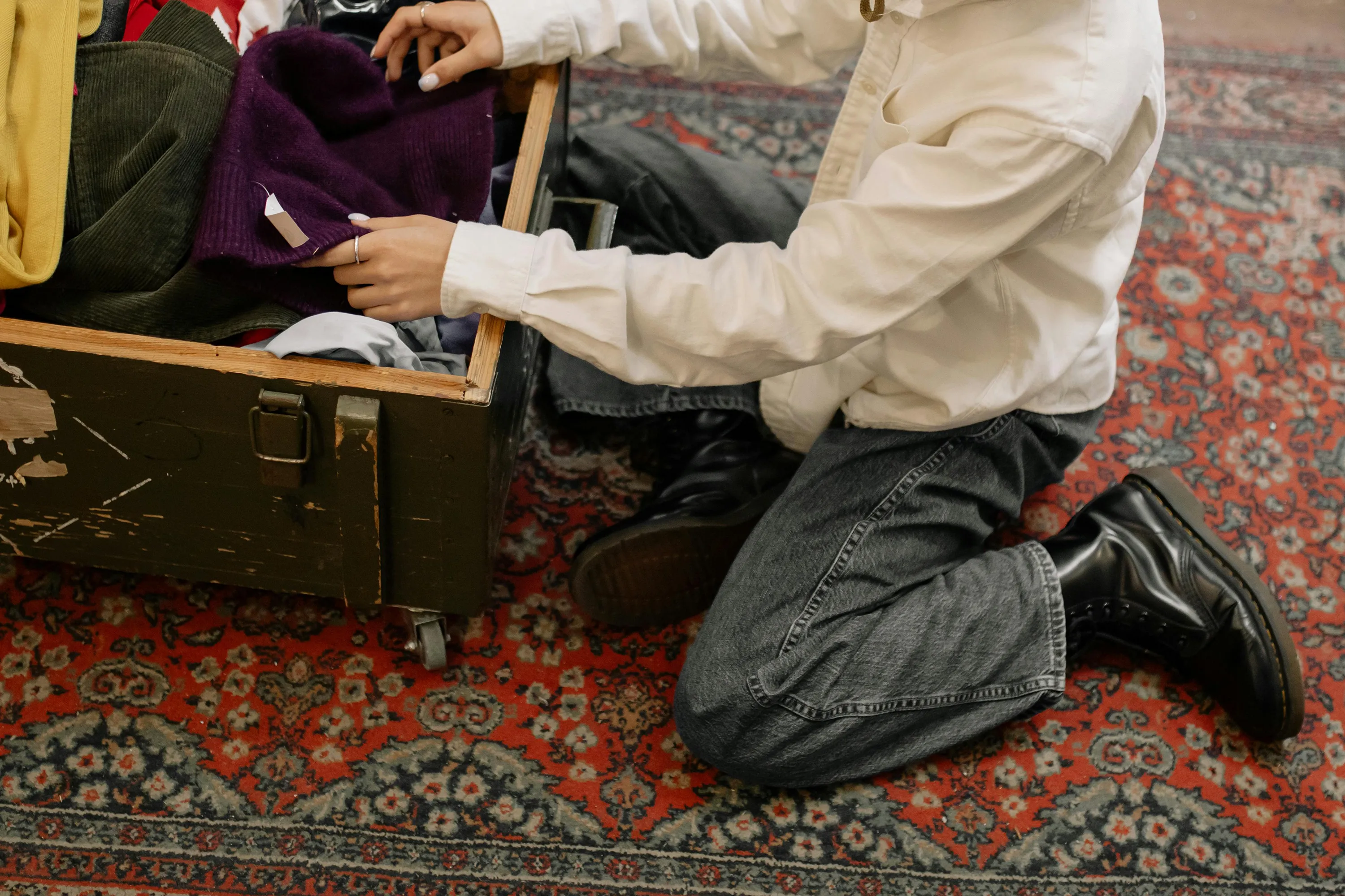 A person kneels on a patterned rug while sorting through clothes in a wooden chest