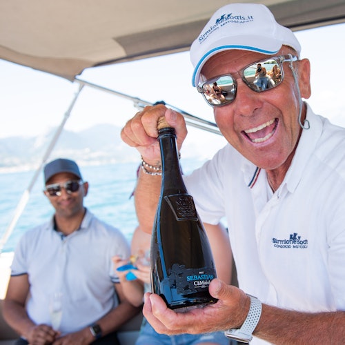 Smiling man in sunglasses and a white polo shirt holds a dark bottle on a sunny boat with other people in the background.