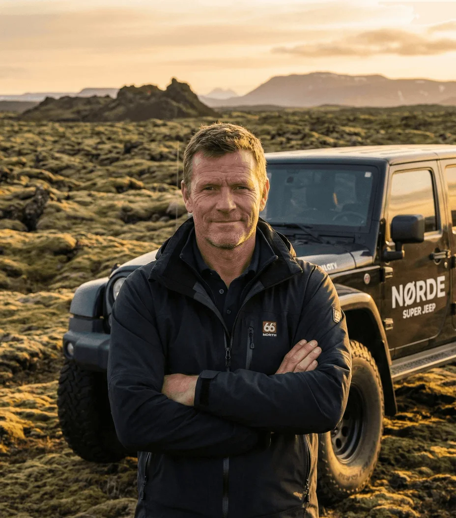 Man in a dark jacket standing with arms crossed in front of a jeep in a mossy field.