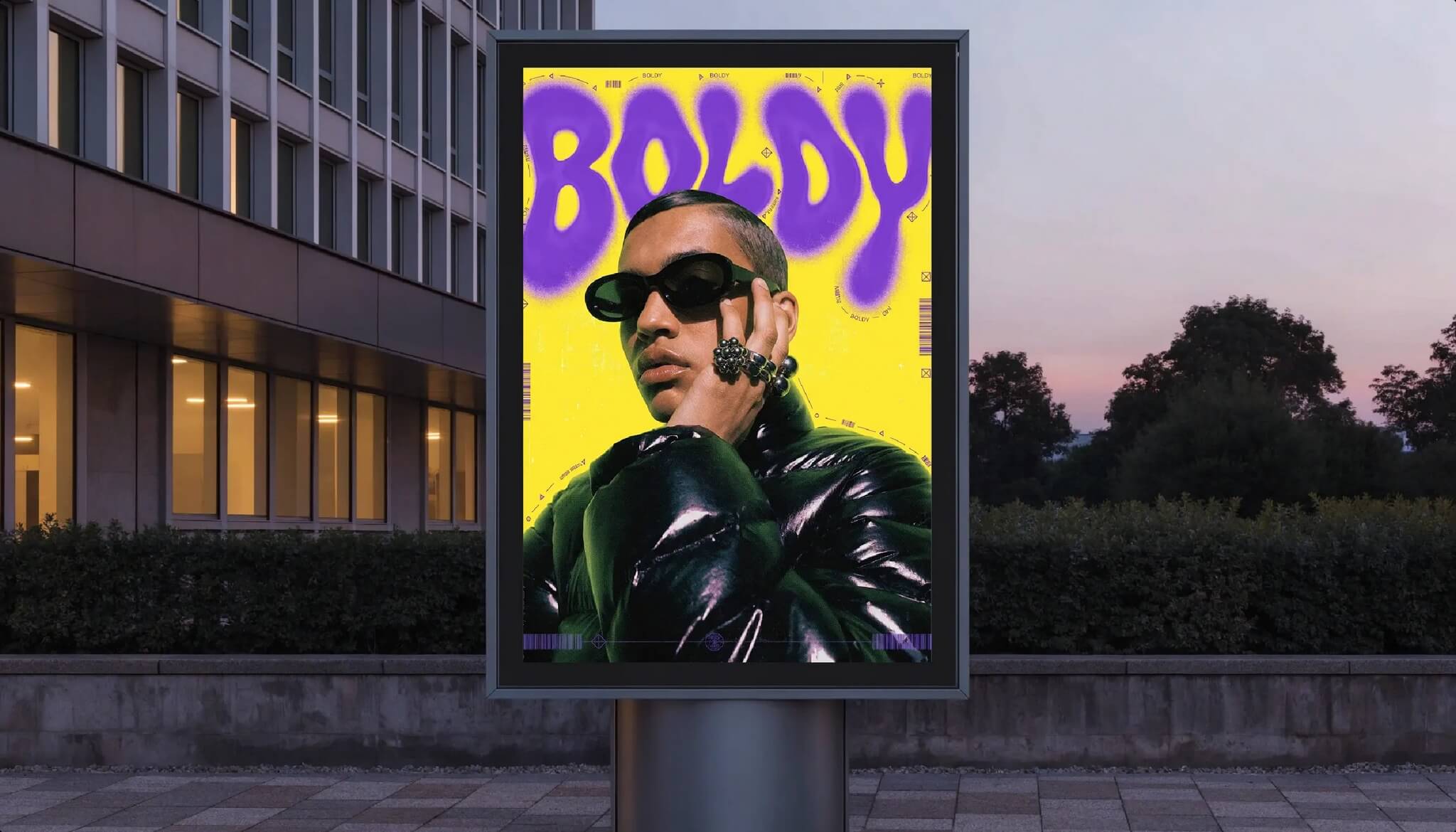 Street billboard at dusk displaying a yellow poster with purple graffiti lettering reading BOLDY over a portrait of a man in sunglasses