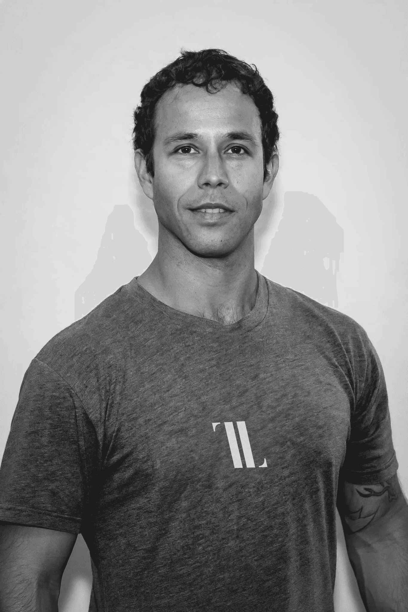 A young person with curly hair smiles at the camera while sitting in front of a laptop against a dark background.