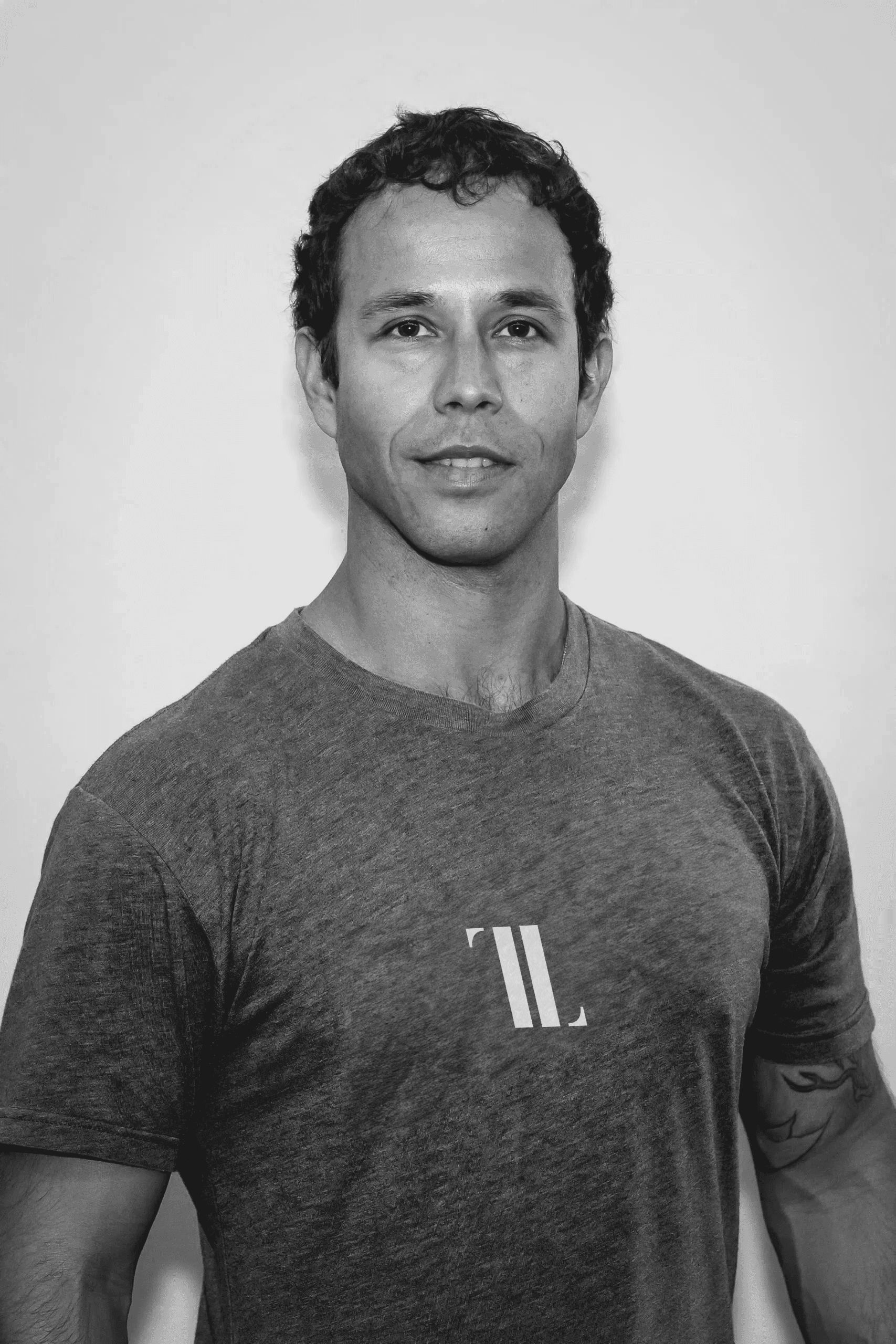 A young person with curly hair smiles at the camera while sitting in front of a laptop against a dark background.