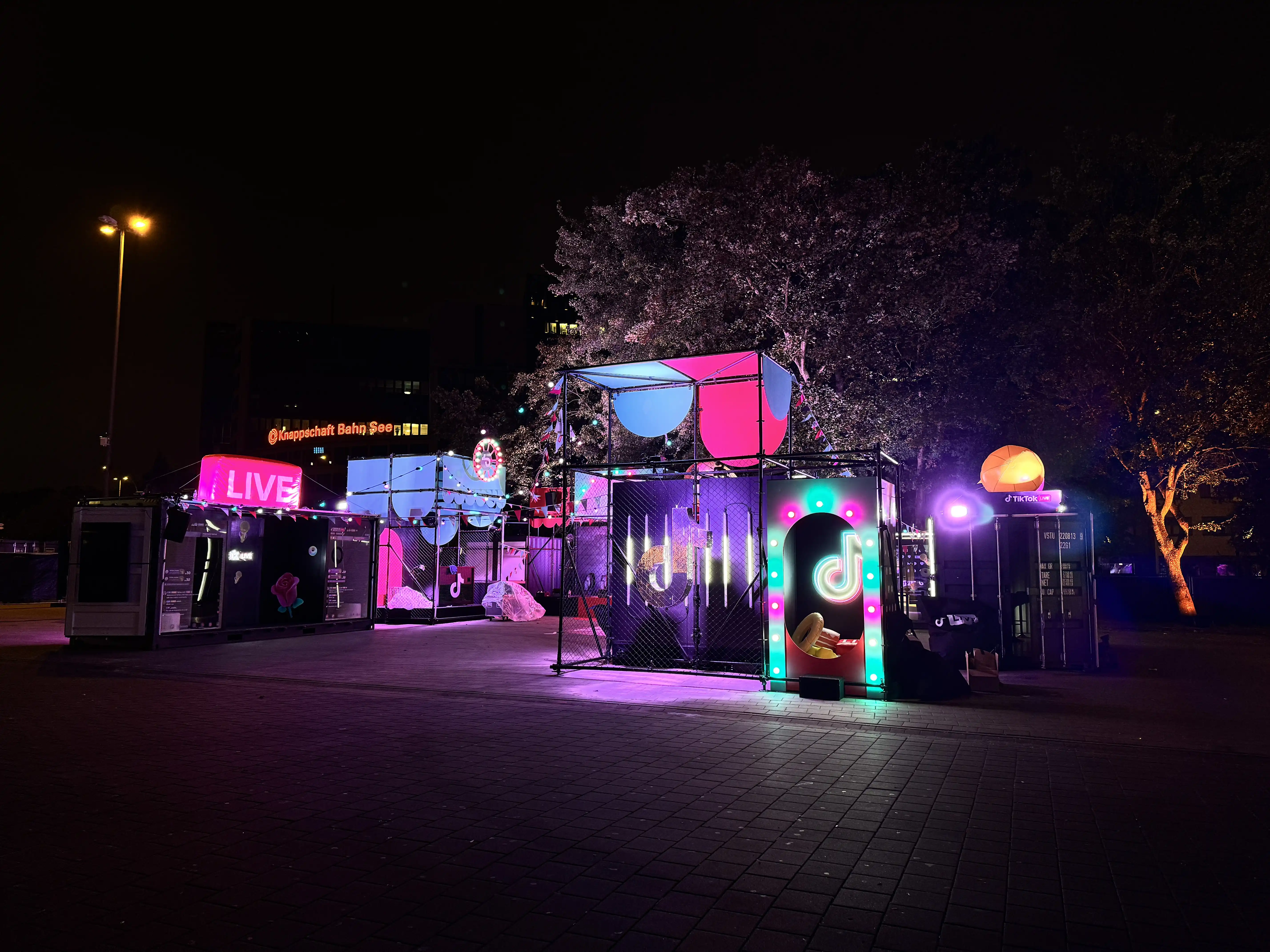 Night scene of the TikTok pop-up with neon arch, Live sign and illuminated canopy shapes.
