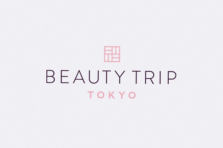 A white iPhone displaying the "BEAUTY TRIP TOKYO" app, featuring an interior view of a store with shelves and displays, along with a large image of a woman's face on the left side of the screen. Below, options to "SIGN IN" and "JOIN" are visible, with the phone positioned on a display stand among additional items.