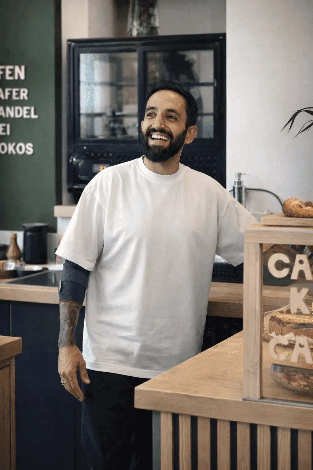 A smiling person in a casual shirt stands next to a wooden counter in a coffee shop.