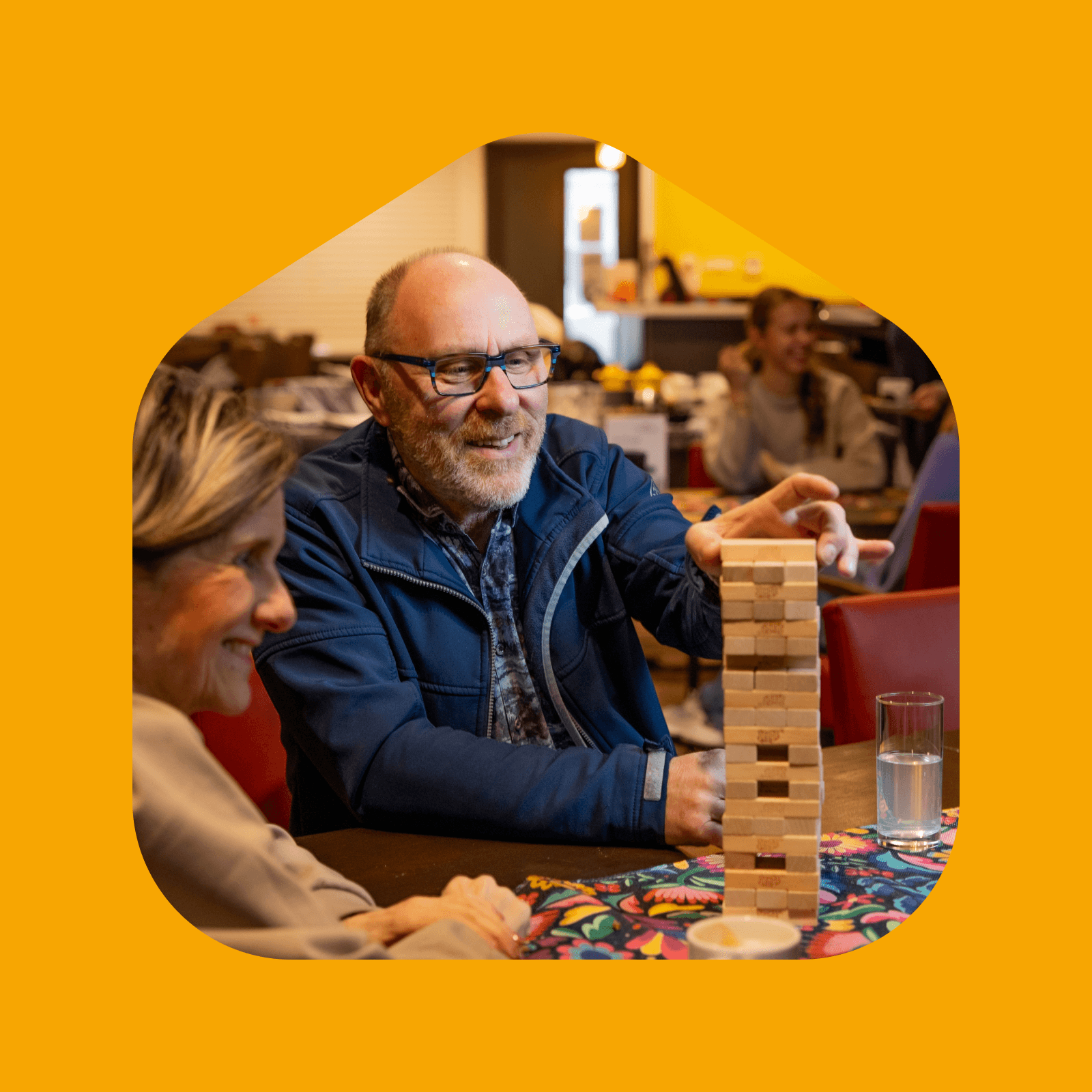 Groep mensen speelt een gezelschapsspel aan tafel tegen een gele achtergrond.