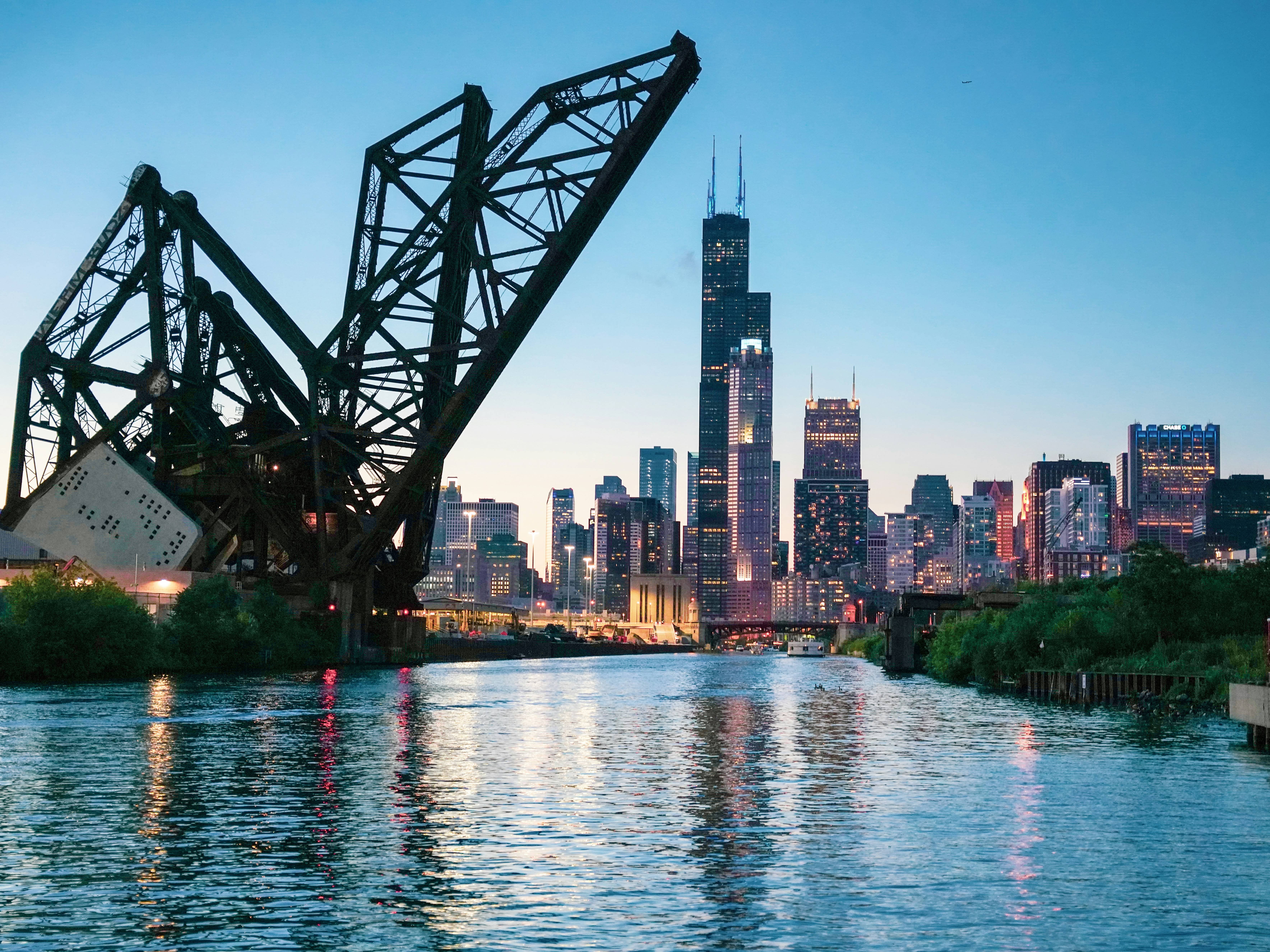 A raised railroad bridge frames the Chicago skyline, including Willis Tower, along the river at dusk.