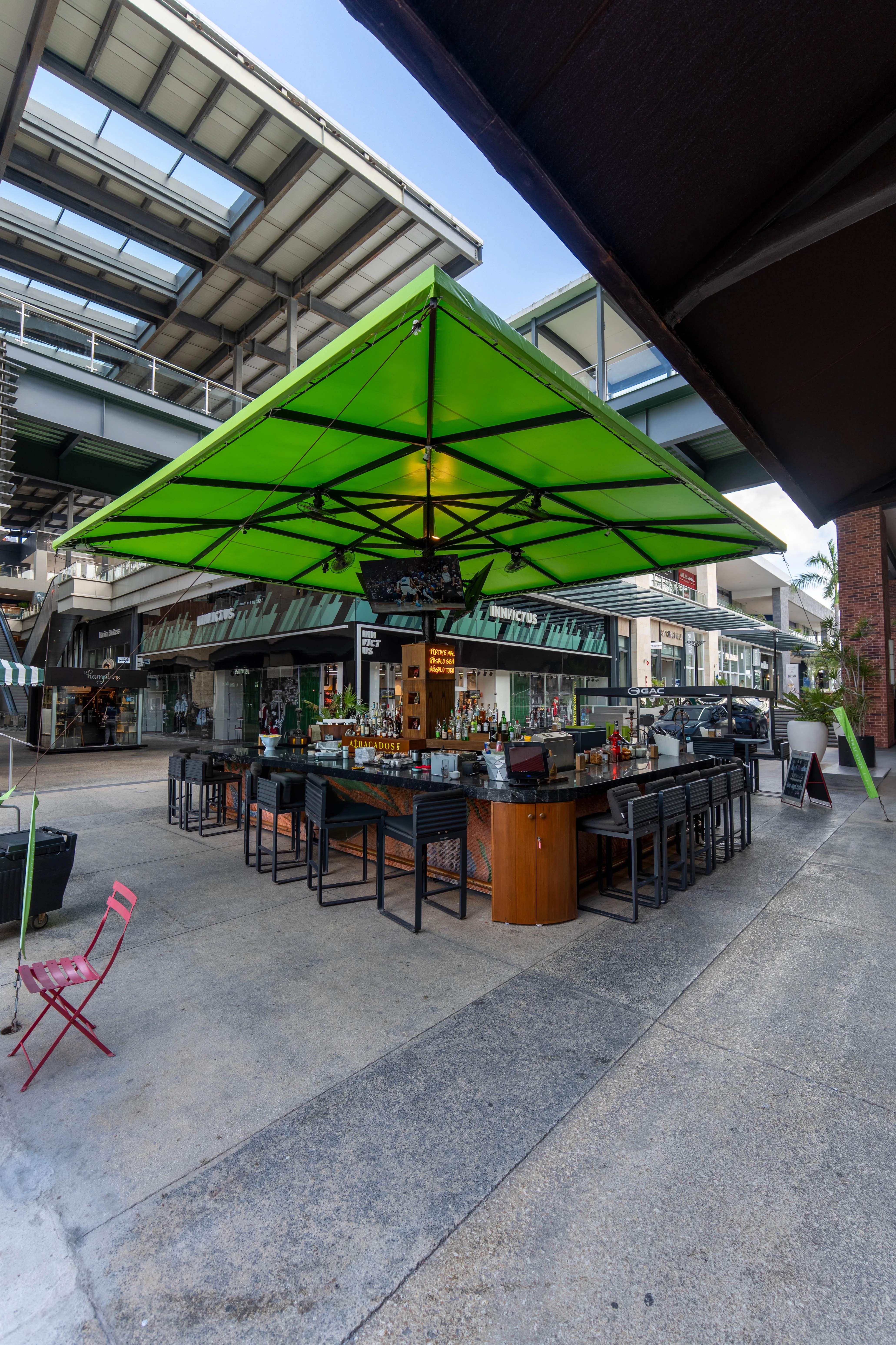 Bar de Atracados con sombrilla verde central, barra oscura, botellas de licor y sillas altas