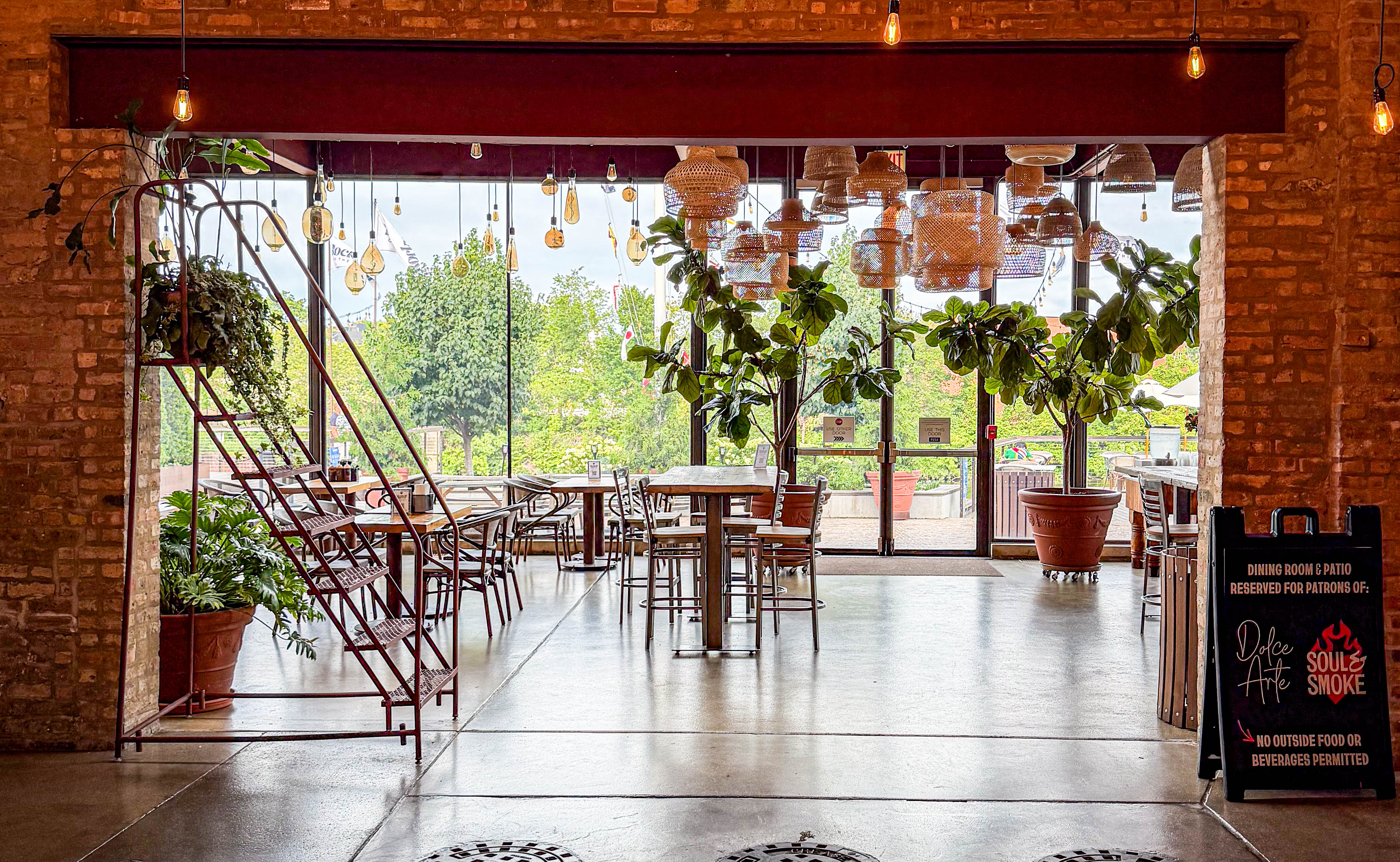 an open exposed brick room with large plants and windows and basket light fixtures with a scissor sign outside