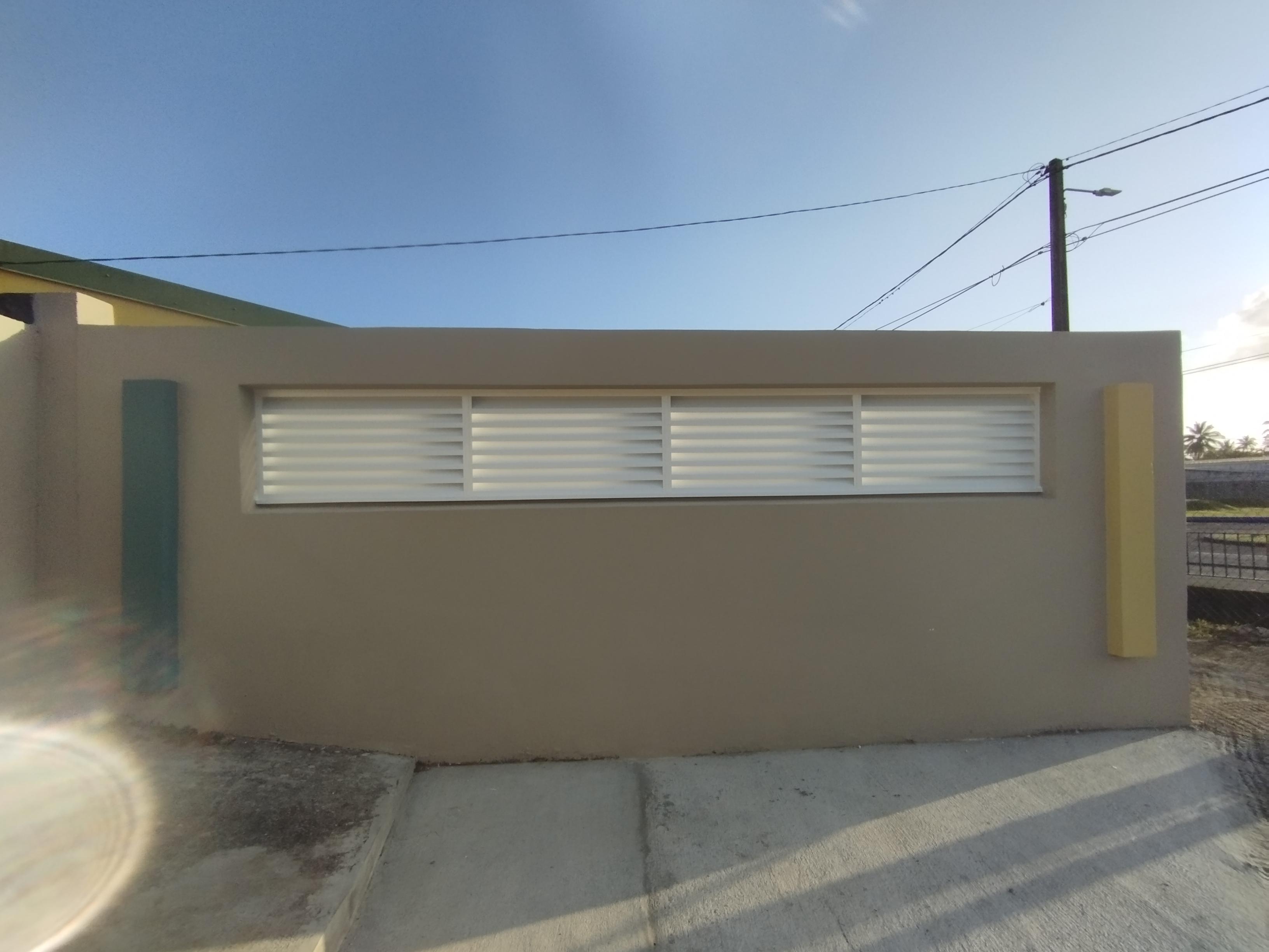 Mur beige avec des bouches d'aération horizontales blanches, encadrées par des accents verticaux bleus et jaunes. Ciel clair et lumineux au-dessus, créant une scène calme et ordonnée.