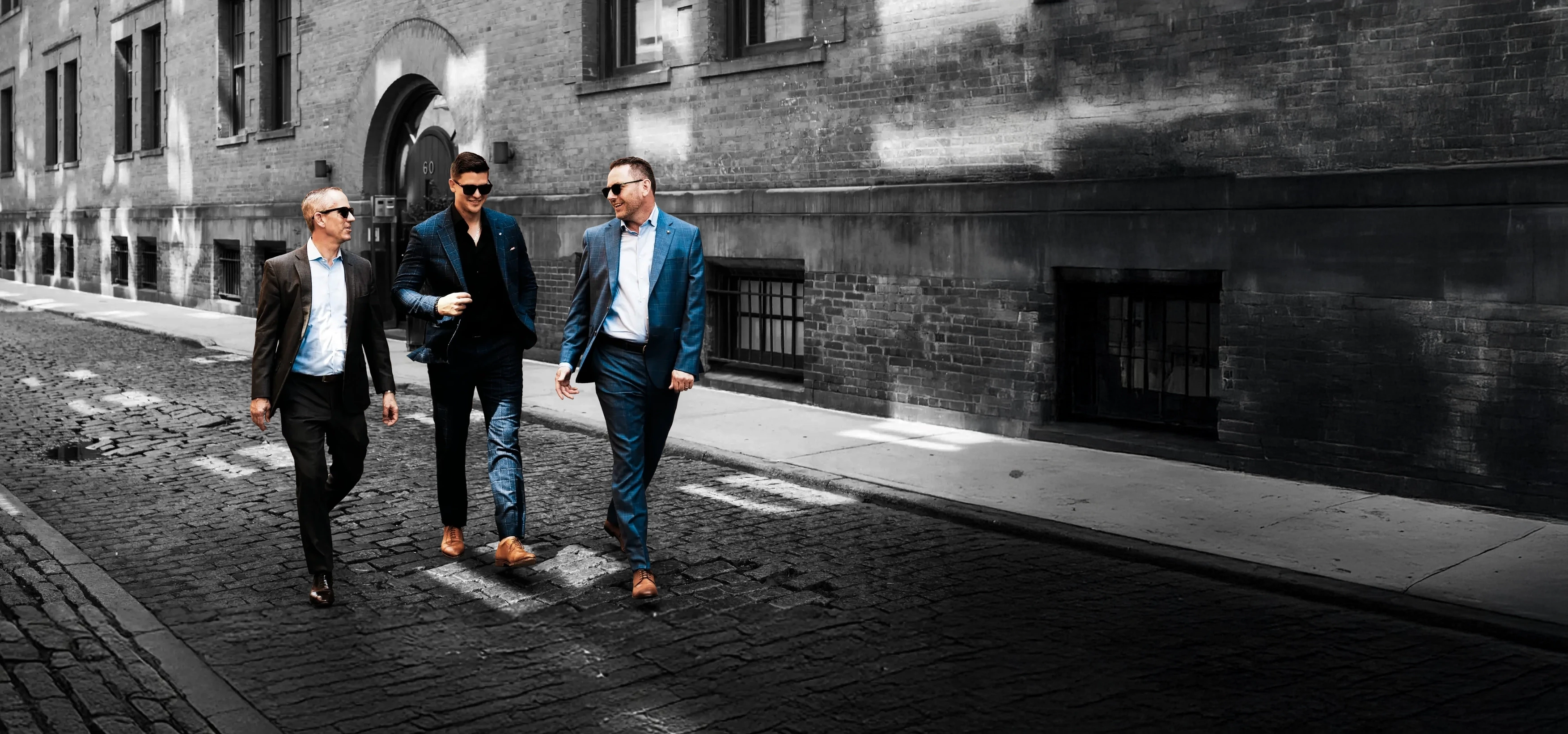 Three business professionals walk and talk along a cobblestone street beside a large building, dressed in smart casual attire.