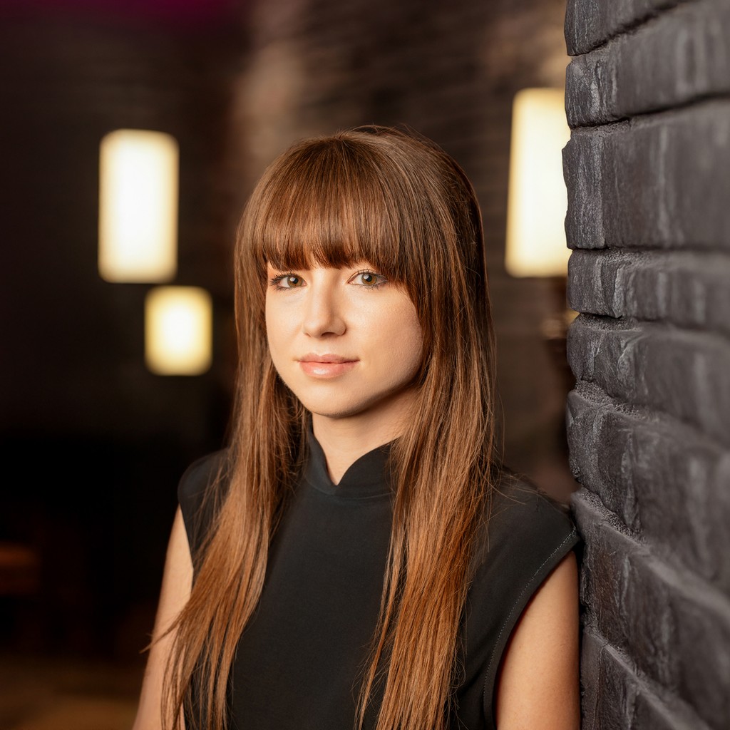 Holly Stewart in a modern office setting, standing against a dark brick wall with warm ambient lighting, wearing a black sleeveless top and straight brown hair with bangs, looking confidently at the camera.