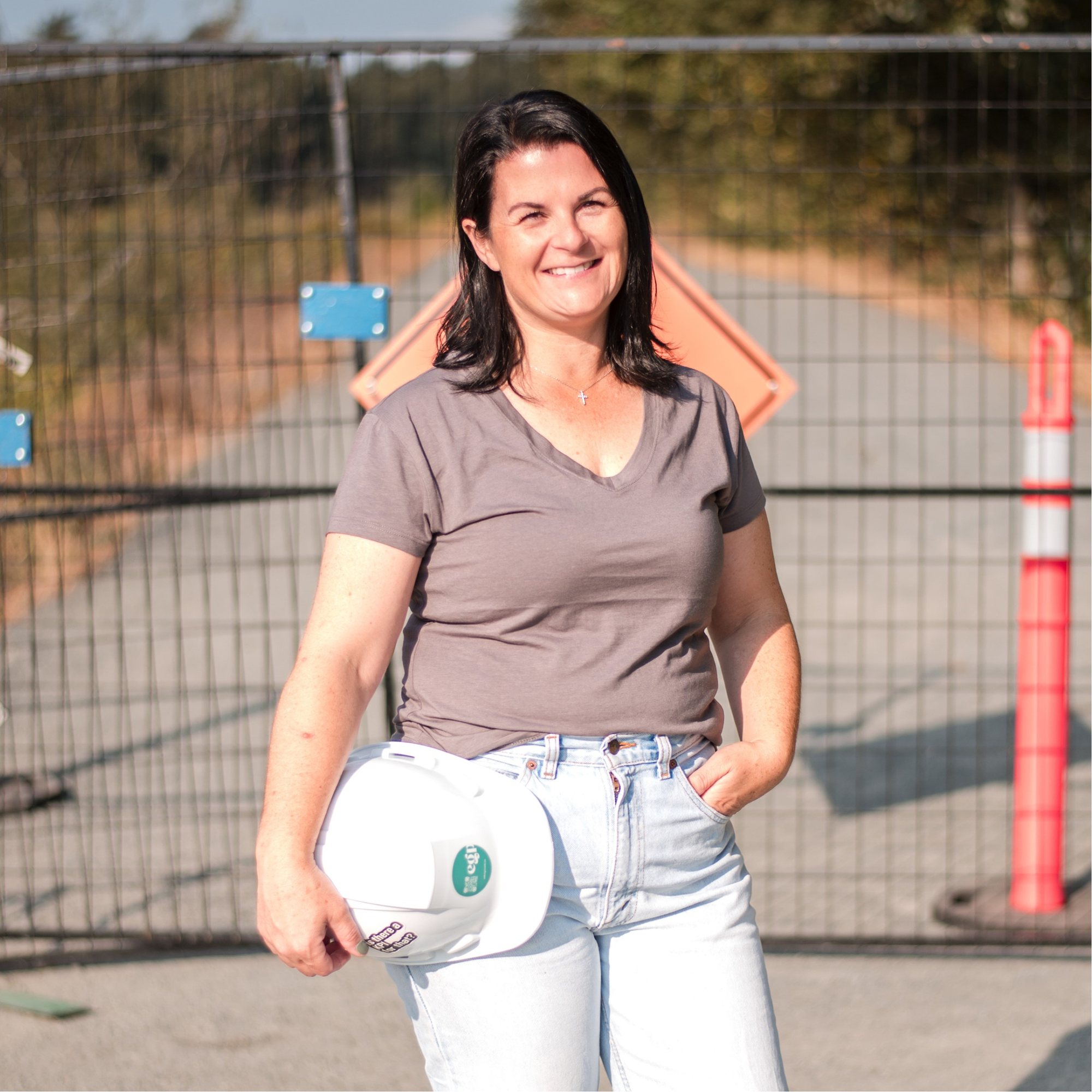 Carey Jones smiling in front of a construction site fence, holding a white hard hat at her side