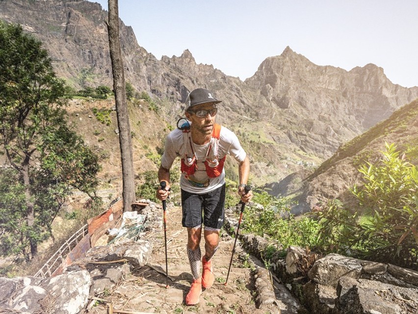 Un coureur grimpe sur un sentier montagneux escarpé, avec des paysages spectaculaires de montagnes autour.