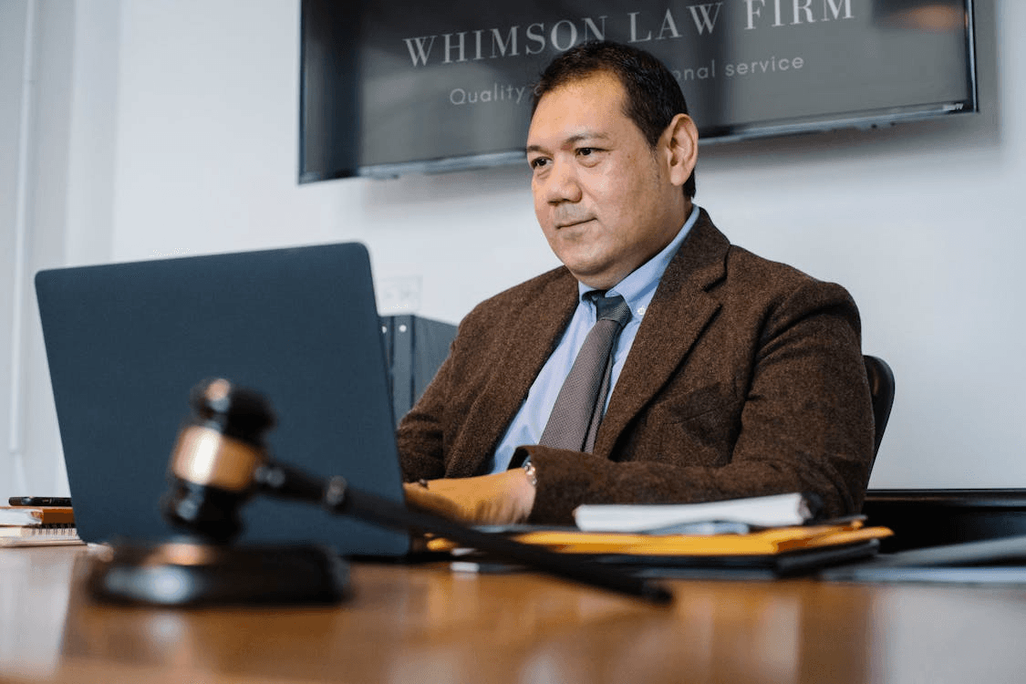 A lawyer in a suit working on a laptop at his desk, with a gavel in the foreground and a law firm sign on the wall behind him.