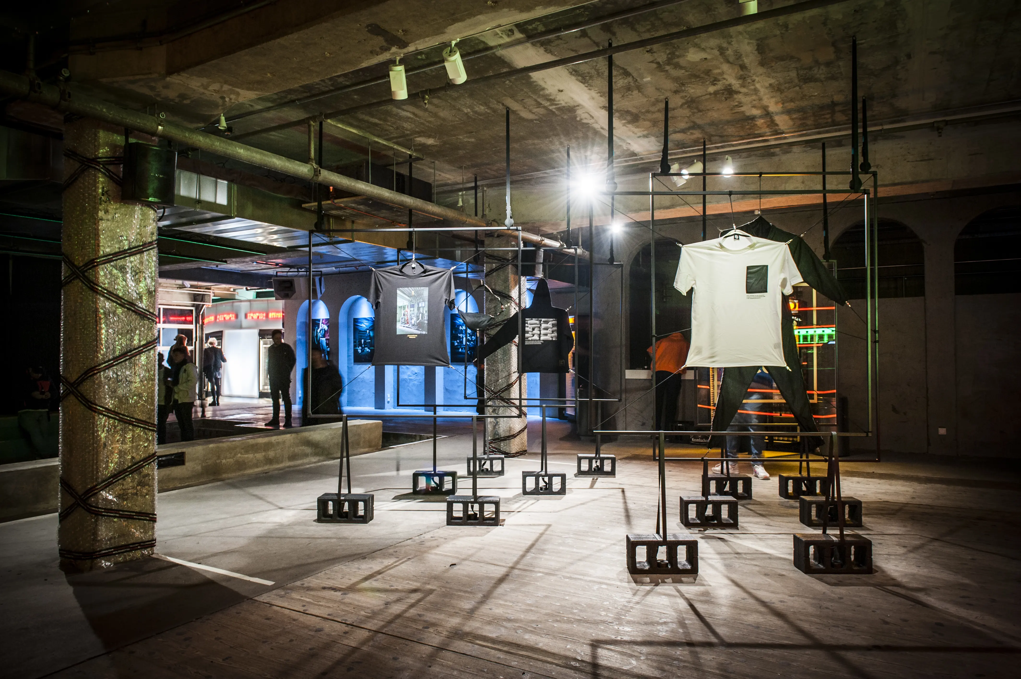 Wide view of the JÄGERMEISTER streetwear installation with shirts hanging from black frames on breeze-block bases in a raw industrial hall.