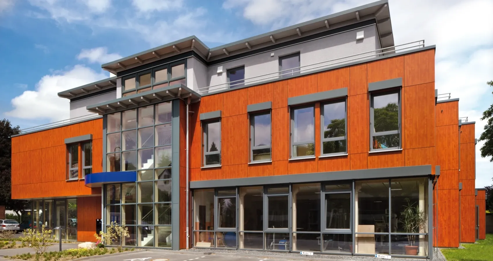 Moderne Bürogebäude mit orangefarbener Holzfassade und großen Glasflächen, im Freien unter blauem Himmel.