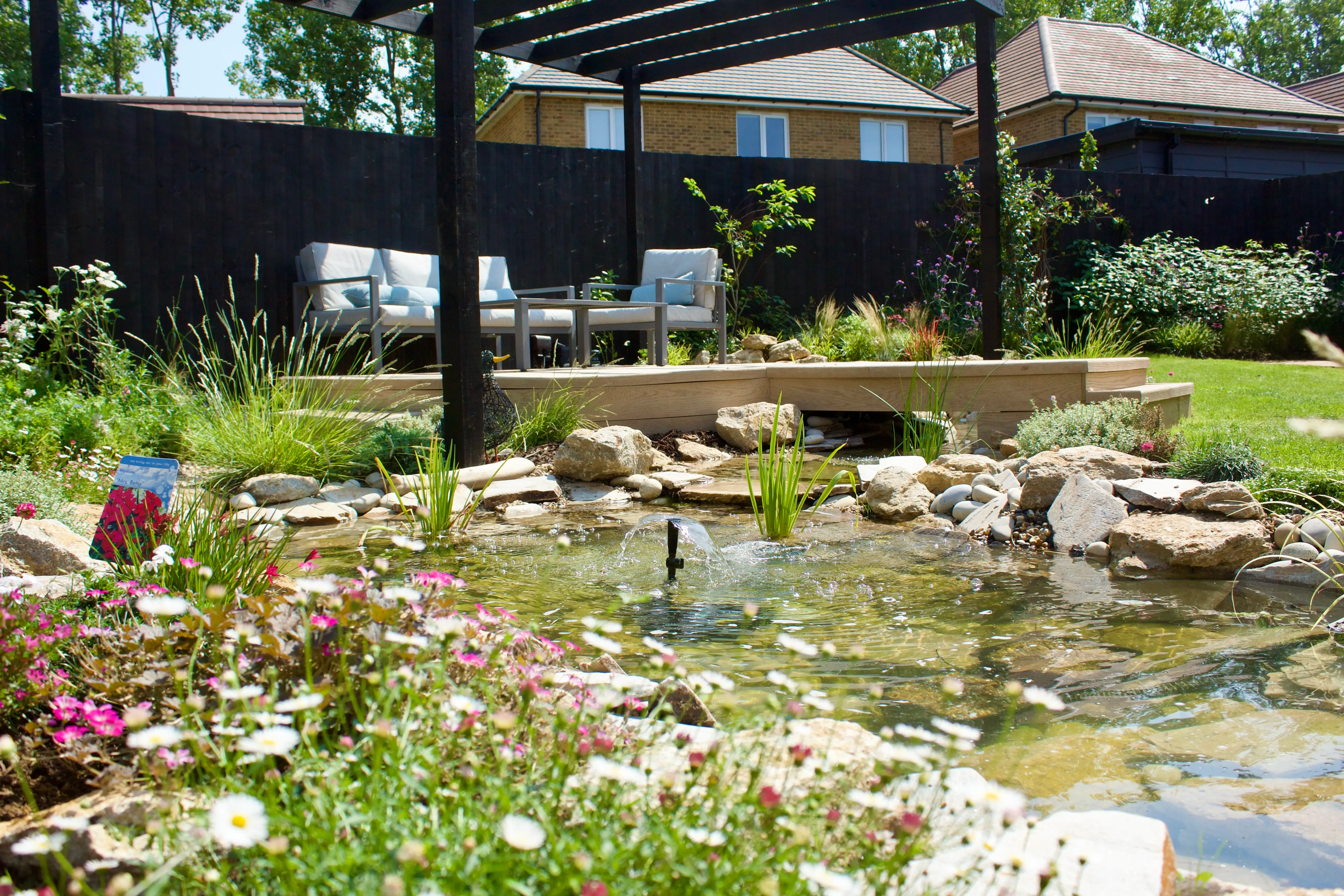 A serene garden pond surrounded by vibrant flowers, rocks, and greenery, with a backdrop of homes.