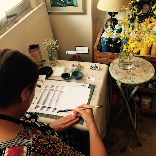 在桌子上绘画的人,周围有各种艺术用品、花瓶中的花朵,以及一张摆放有台灯和更多用品的桌子。