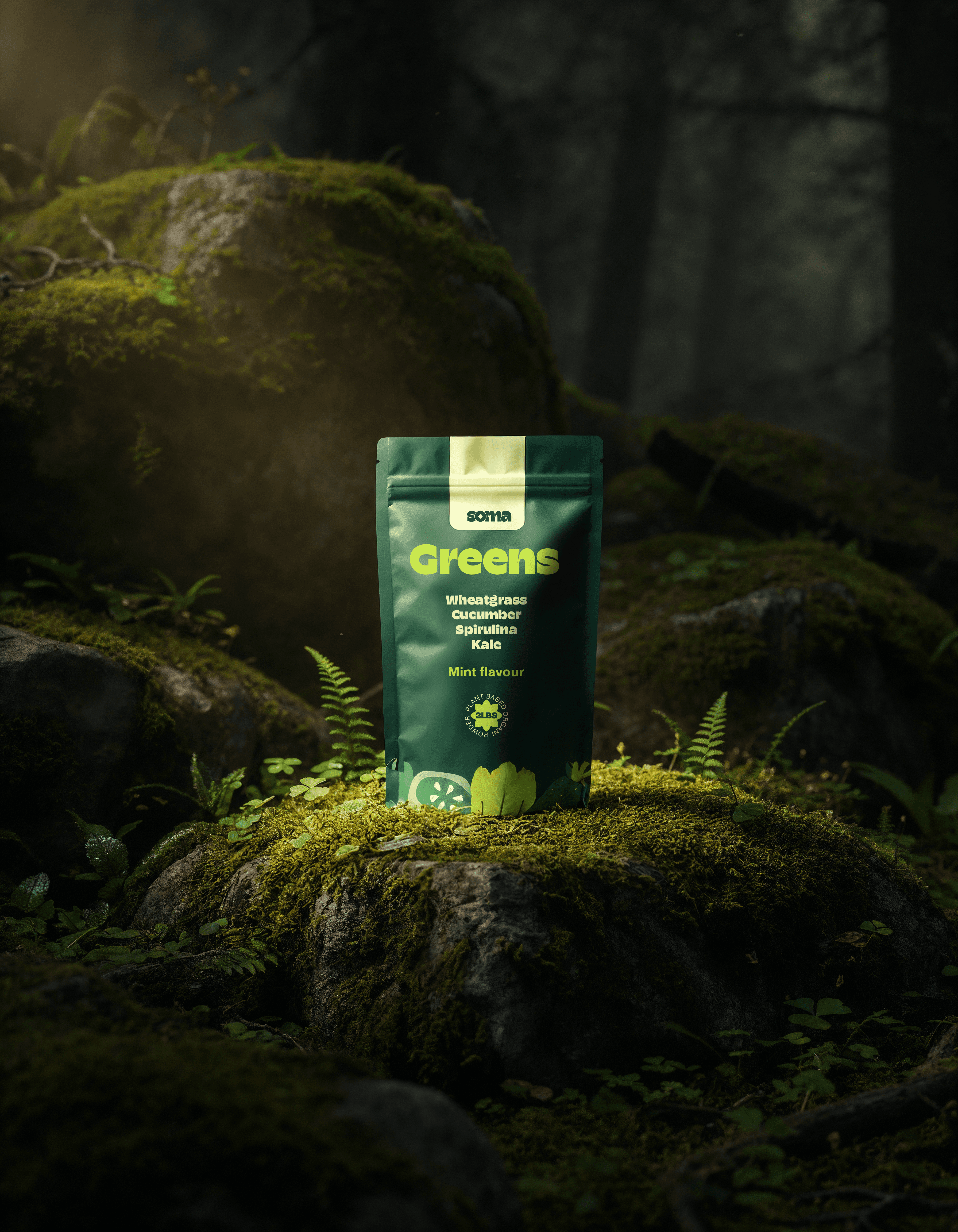 A dark green matte stand-up pouch for "SOMA Greens" supplement, featuring lime green typography. The packaging is placed on a mossy rock in a moody, sun-drenched forest setting with ferns and organic light leaks.