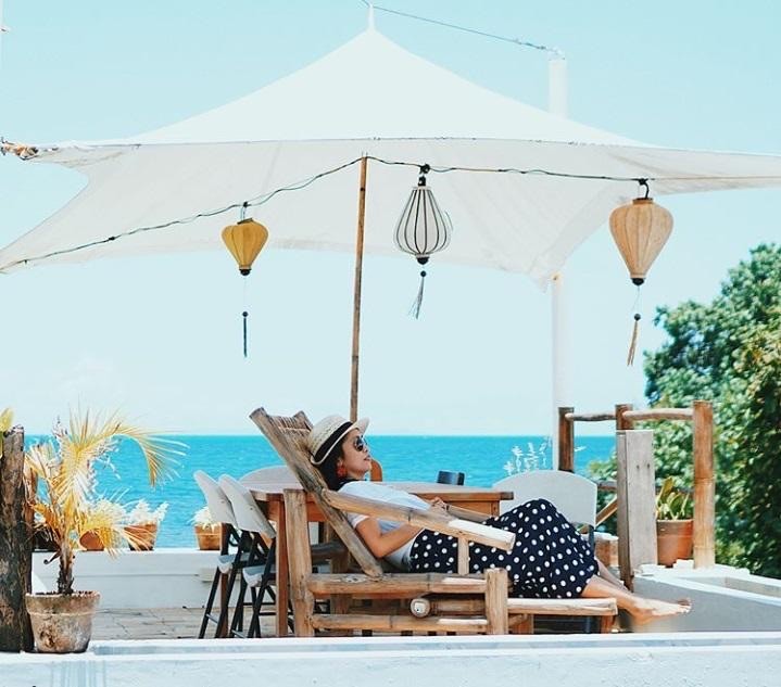 A woman relaxing in a wooden lounge chair under a white canopy with hanging lanterns, overlooking the blue ocean.