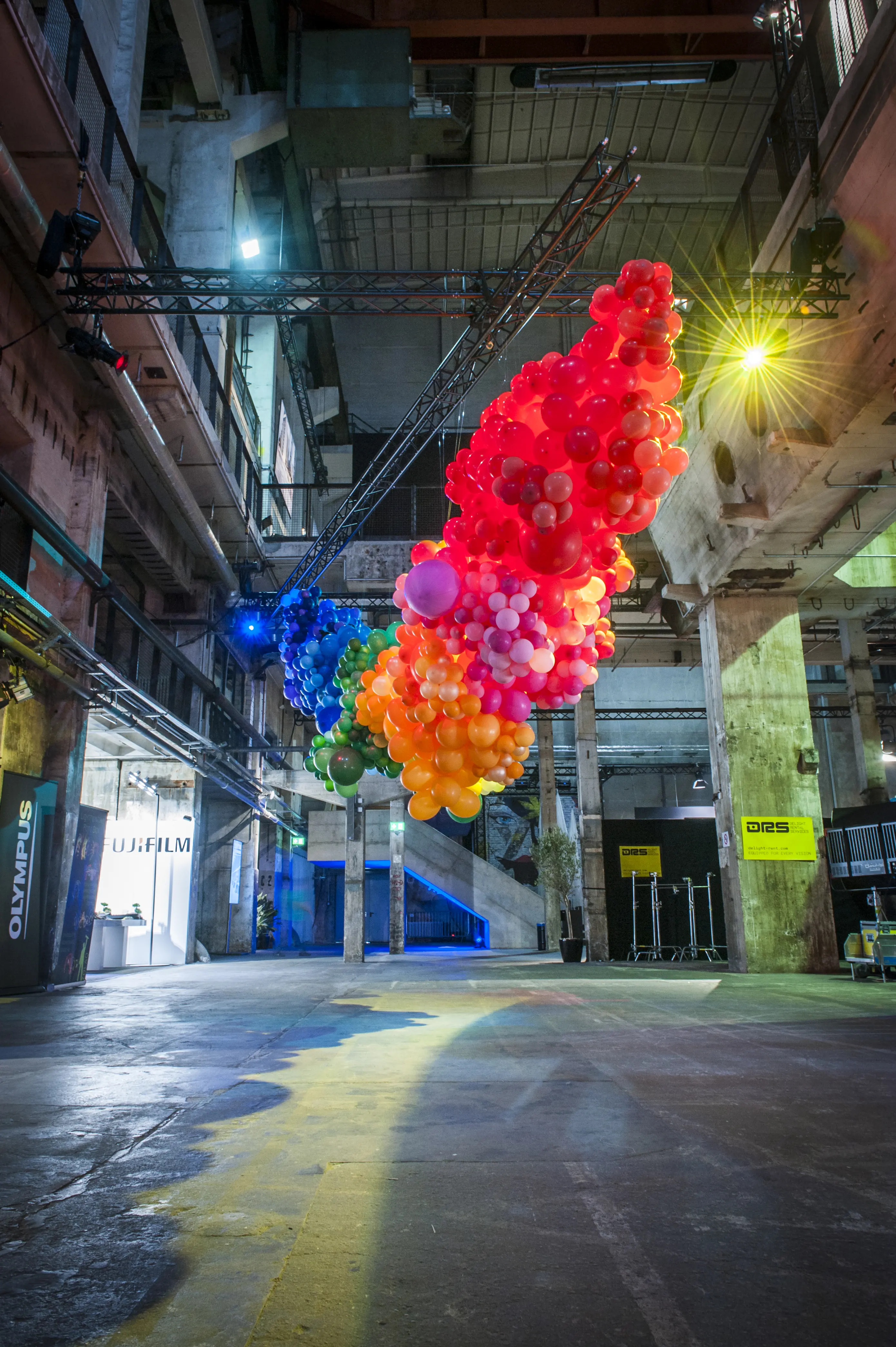 Rainbow-colored cloud of balloons floating diagonally through the industrial hall at BERLIN PHOTOWEEK.