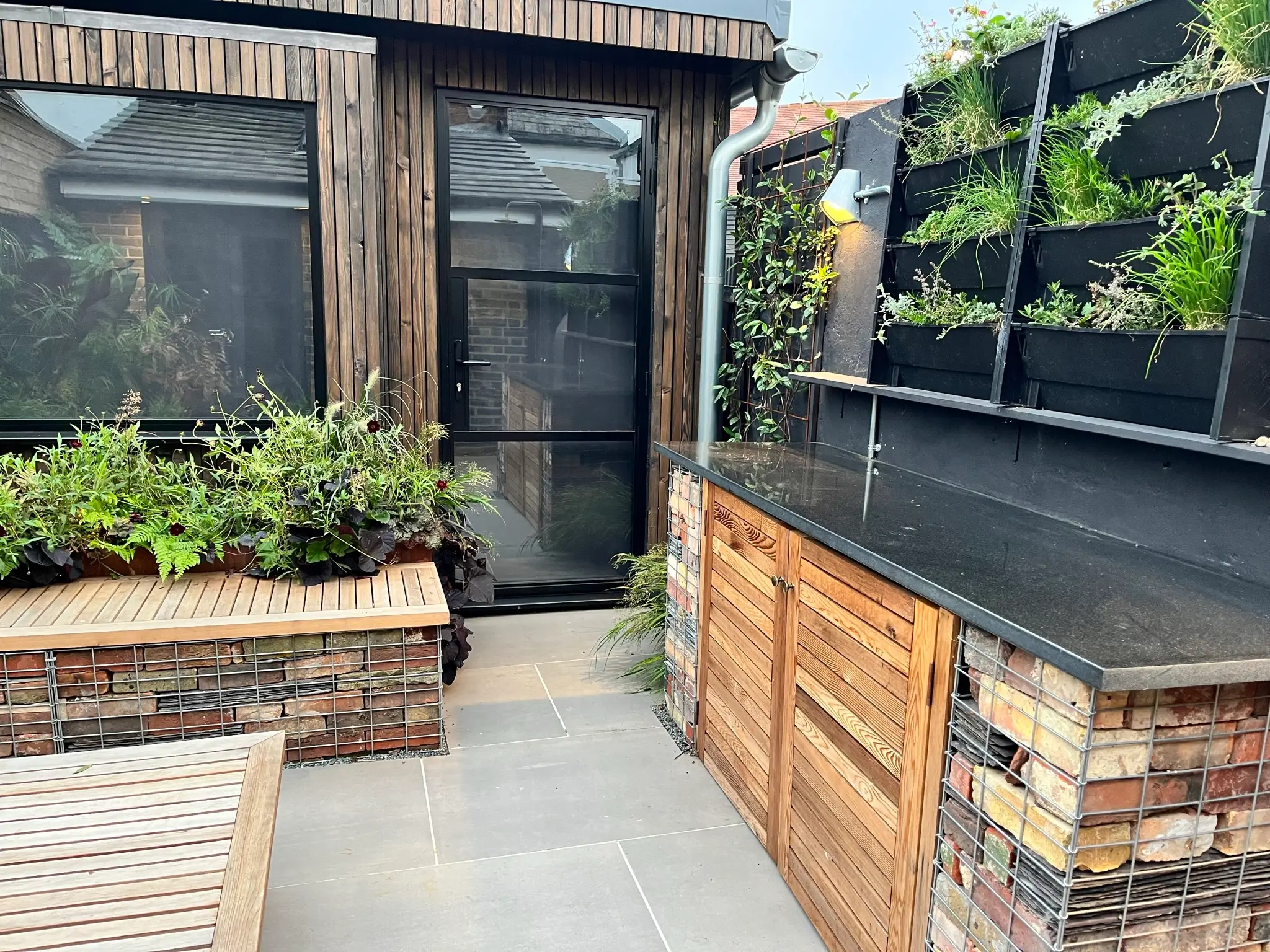 A cozy outdoor space featuring a wood and stone bench, green plants, and a glass door leading inside.