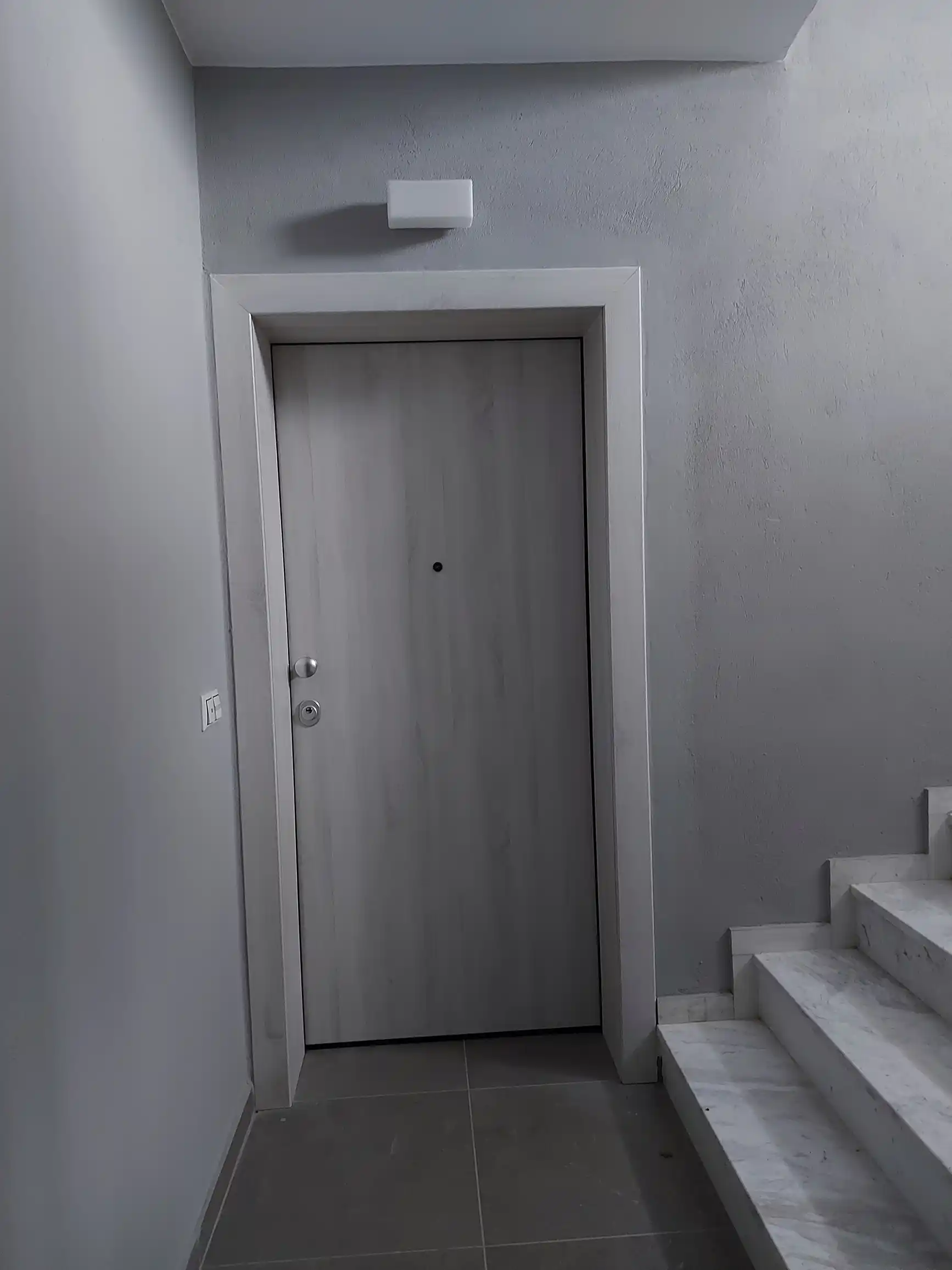 Μοντέρνα ανοιχτόχρωμη πόρτα εισόδου διαμερίσματος σε γκρι κλιμακοστάσιο με μαρμάρινη σκάλα.