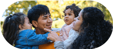 A happy couple playing with their two young daughters outdoors. The father holds one daughter and the mother holds the baby, who is looking intently at her mother